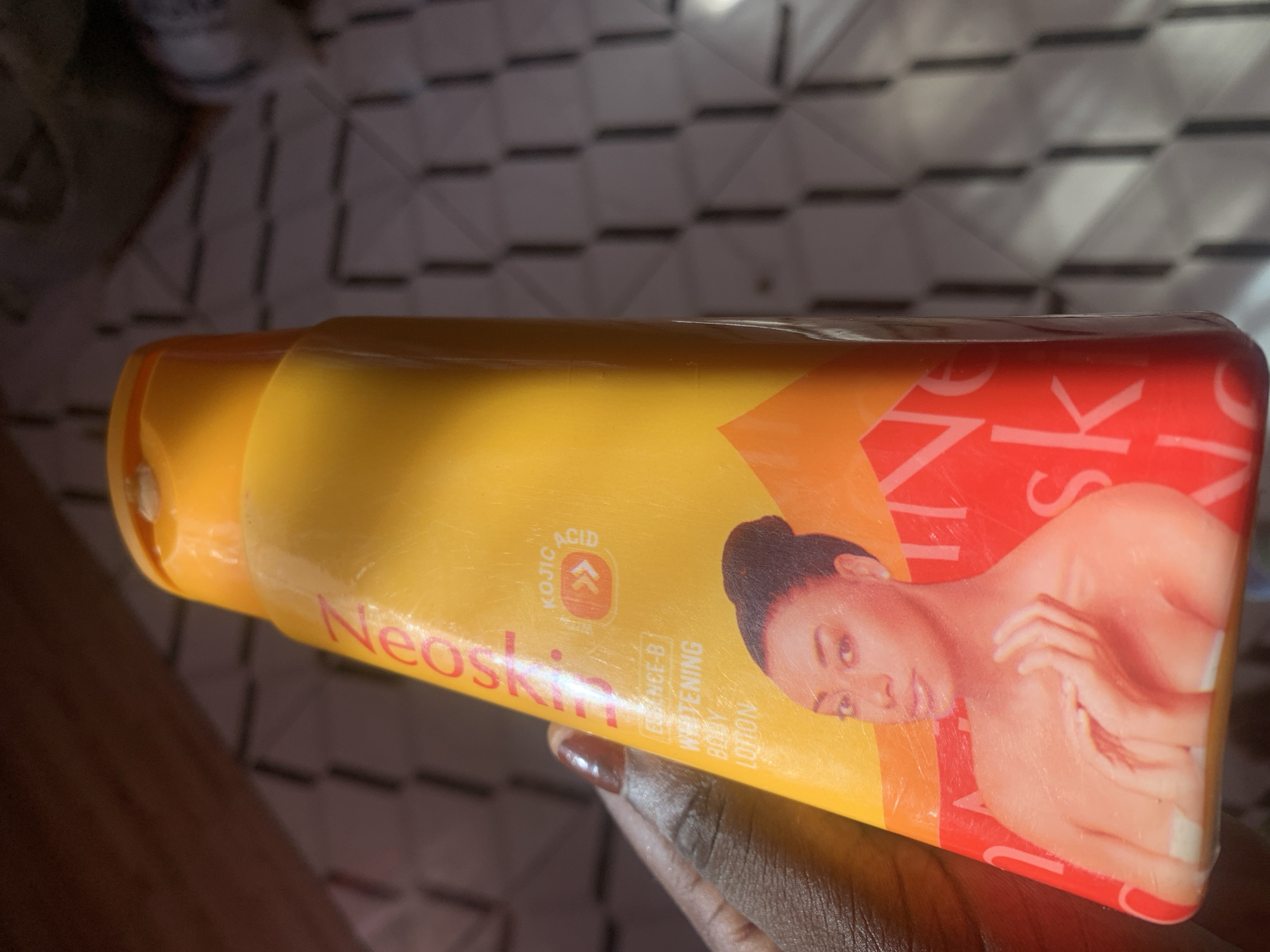 A close-up shot shows a person's hand holding a bottle of Neoskin Essence-B Whitening Body Lotion. The bottle is primarily yellow and orange with a picture of a woman's face and upper torso. Text on the bottle includes "Neoskin," "KOJIC ACID," and "ESSENCE-B WHITENING BODY LOTION." The background is blurred, showing a patterned surface and some wooden elements. The overall lighting suggests an indoor setting. There is no indication of Jauro Yinu, Nigeria in the image itself.