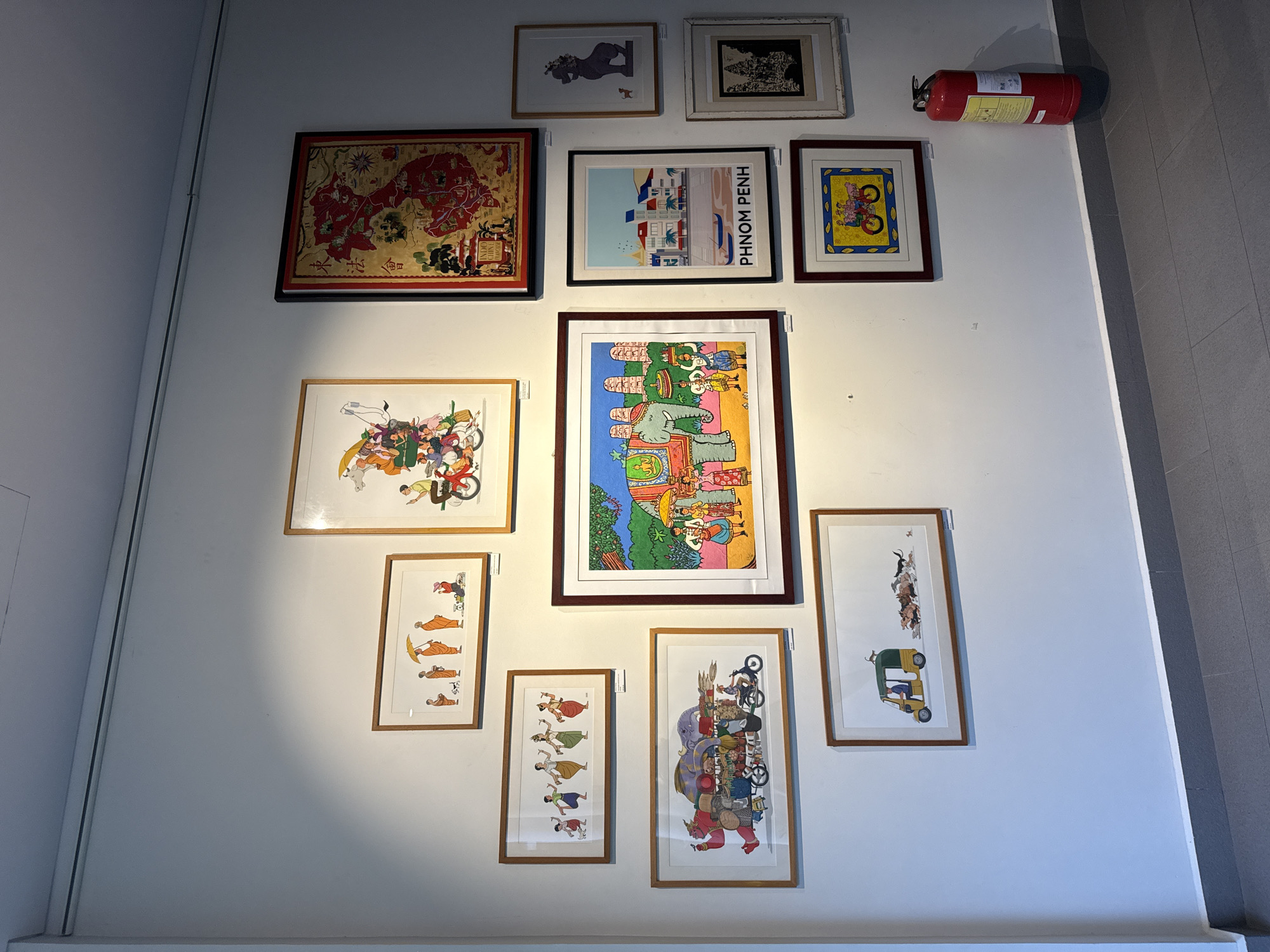 The image depicts an art exhibition in Phnom Penh, Cambodia, possibly within the Khan Daun Penh district. The scene is dominated by a white wall displaying numerous framed artworks. The collection includes various styles and subjects, ranging from colorful, cartoonish illustrations of people and animals to more traditional-looking maps and detailed etchings.
In the center-left of the frame, a large, horizontal artwork showcases a procession of monks walking with umbrellas, followed by other figures and a dog. Below this, another horizontal piece depicts several women in traditional attire dancing. Further down, a large, framed drawing features an elephant carrying a heavy load, with a person riding a scooter beside it. To the right of these, a vibrant, square artwork depicts people on bicycles and a tuk-tuk.
To the right, a prominent, vertically oriented framed artwork displays a red map with intricate details, likely of Cambodia, with gold lettering. Below this, a framed print depicts a stylized silhouette of an animal with a small dog in front of it. Further down, a smaller framed artwork shows what appears to be a temple or ancient structure. Another framed print presents a cityscape with buildings and boats, labeled "PHNOM PENH."
In the foreground, at the bottom right of the image, a bright red fire extinguisher stands upright against the wall, its presence suggesting a public venue. The floor is tiled with dark grey tiles. The lighting in the room appears to be artificial, casting subtle shadows on the wall. The overall impression is that of a curated display of art within a gallery or exhibition space in Phnom Penh.