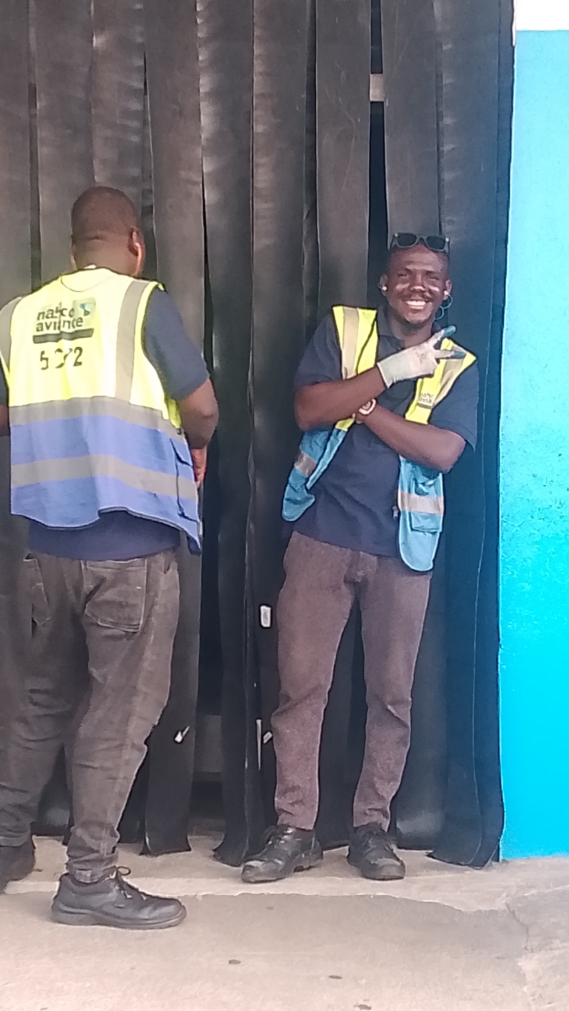 Two men are standing in front of a black curtain or backdrop next to a blue wall. The man on the left is facing away from the camera, wearing a yellow and blue safety vest, a dark blue shirt, and grey pants. The man on the right is facing the camera, smiling, with sunglasses on his head, a blue and yellow safety vest, a dark blue shirt, and brown pants. He is wearing a white glove and giving a peace sign. The location is Orerokpe, Nigeria.