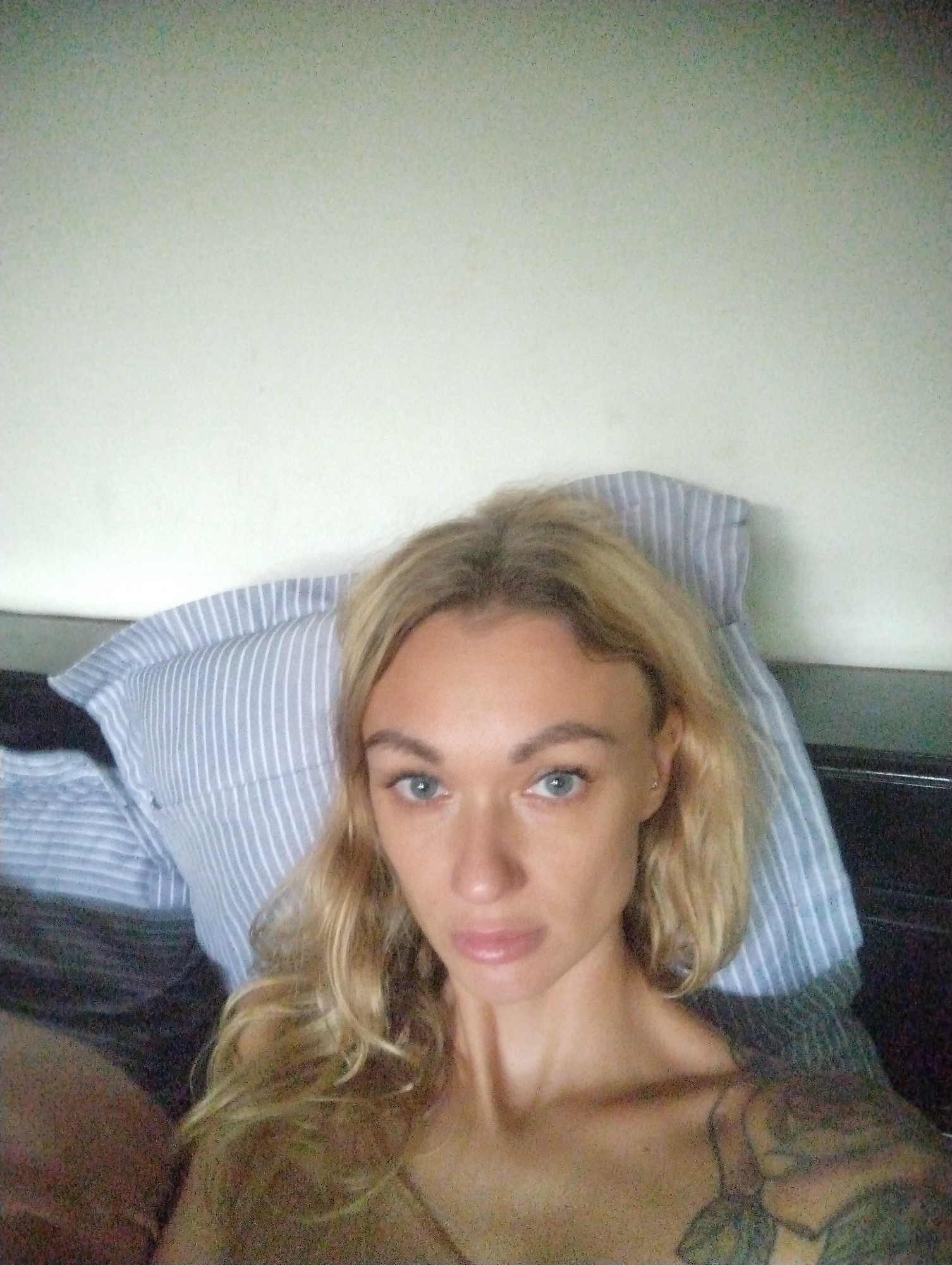 A woman with blonde hair, blue eyes, and fair skin is in a bedroom. She appears to be lying in bed with her head resting on a pillow with blue and white stripes. She has a tattoo on her upper chest and shoulder. The wall behind the bed is white. The location of the image is possibly in the city of Mũi Né, Vietnam.