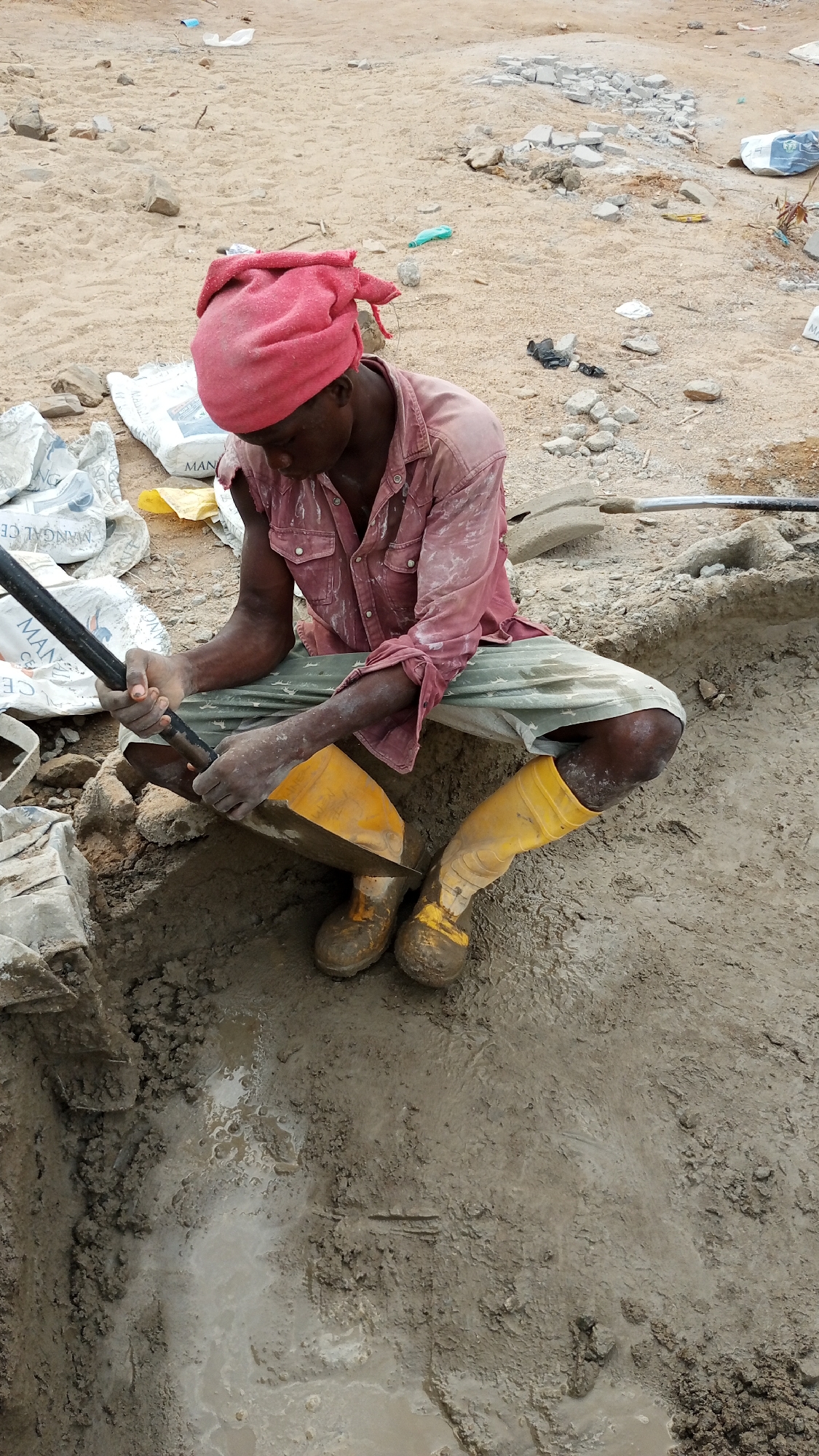 A man squats in a muddy pit in Zango-ndaji, Nigeria. He wears a red head wrap, a pink long-sleeved shirt, khaki shorts, and yellow rubber boots. He holds a shovel and uses a hand tool to mix the mud in the pit. Bags of cement and debris are scattered on the ground around the pit. The environment is dusty and appears to be a construction site.