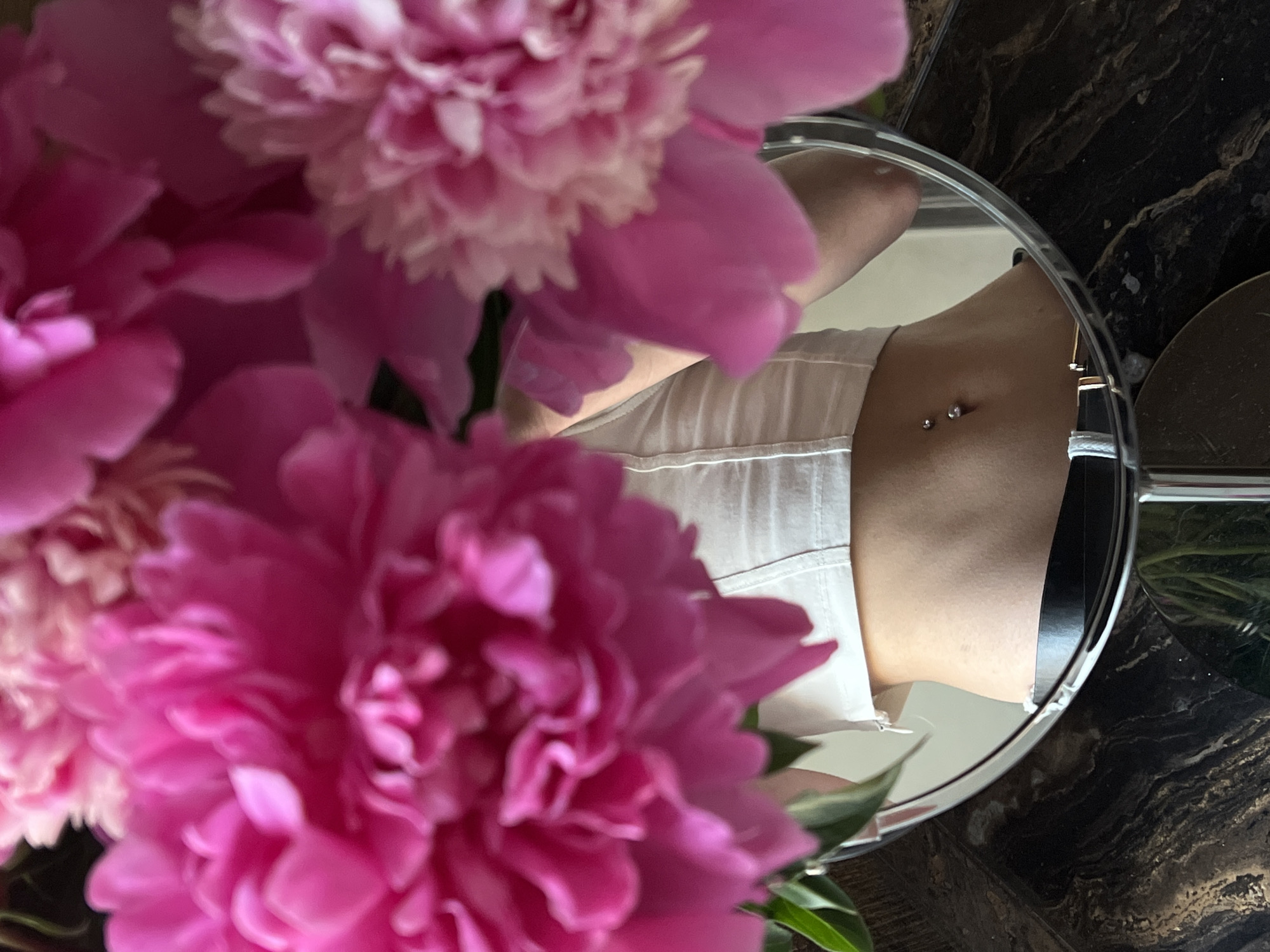 A close-up shot captures a reflection in a round mirror of a person's midriff, adorned with a belly button piercing. The mirror is surrounded by vibrant pink peonies, some in sharp focus and others blurred. The background visible in the mirror suggests an indoor setting, possibly a home or studio, with a dark, marbled surface beneath the mirror. The image does not contain any information about the location in Ternopil, Ukraine.