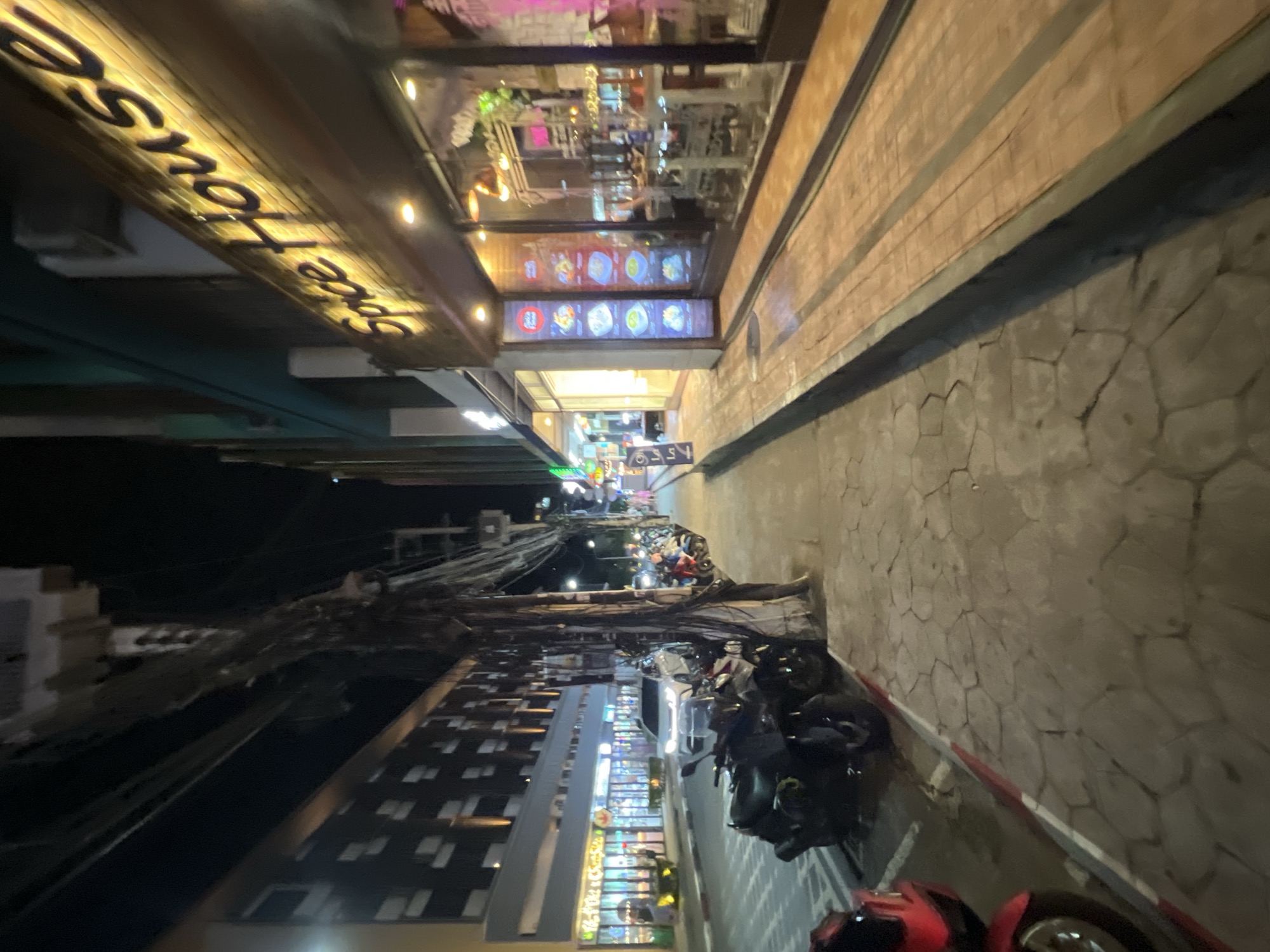 The image depicts a street scene in Karon, Thailand, at night. The perspective is low, looking upwards and diagonally along a street lined with buildings and parked motorcycles.
On the left side of the street, a building features a prominent illuminated sign that reads "Spice House". Above this sign, the facade of the building has glass windows reflecting the interior, which appears to be a brightly lit establishment, possibly a restaurant or shop with colorful displays. Further down the street, more buildings with illuminated signs and windows are visible. A dense network of electrical wires hangs above this side of the street.
The right side of the street is dominated by a sidewalk paved with irregular, light-colored stones. A dark band runs along the edge of the sidewalk. A row of parked motorcycles is visible along the sidewalk, suggesting a popular area with nightlife or dining options. The lighting is artificial, with streetlights and signs illuminating the scene.
The overall atmosphere is that of a bustling commercial street at night. The time of day is clearly nighttime due to the artificial lighting and the darkness in the background. The weather appears to be clear, as there are no indications of rain or adverse conditions. The presence of the "Spice House" sign and the general architectural style suggest a location in Southeast Asia, consistent with Karon, Thailand. There are no people clearly visible in the image, but the presence of parked vehicles indicates activity in the area.