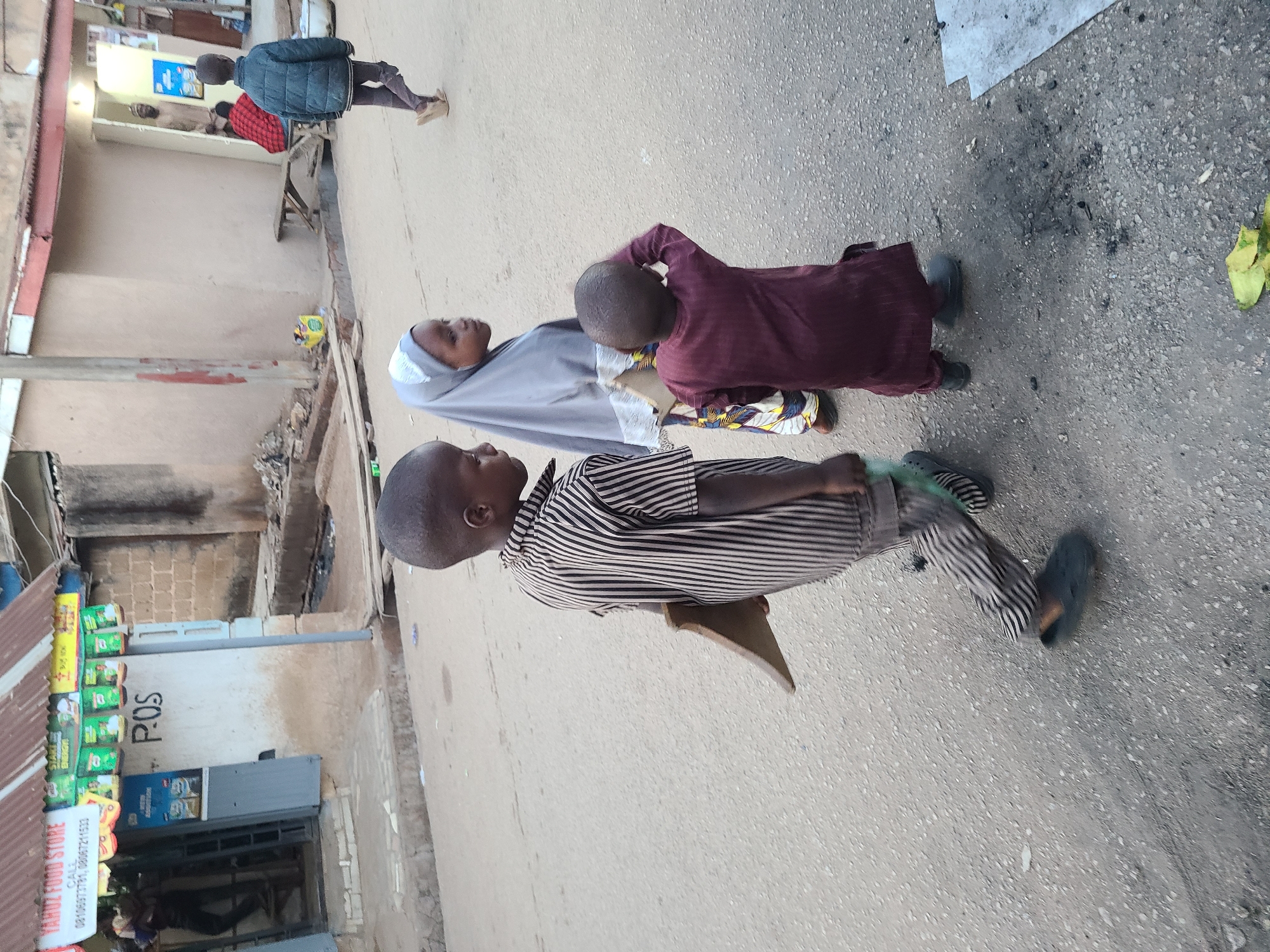 The image, rotated correctly, captures a daytime street scene featuring several individuals and roadside structures. In the foreground, three young children are present. One child, wearing a striped shirt and pants, is seated on a low wooden stool or plank, looking towards the left. To their right, another child in a light grey head covering (hijab) and patterned dress stands, while a third child in a maroon long-sleeved top and a multi-colored patterned skirt stands beside them, seemingly looking towards the child in grey.
Further down the street, a person in a dark blue jacket and dark pants walks away from the viewer. Inside an open doorway of a building on the left, another individual in a red and black checkered shirt is visible.
The ground is a paved surface, speckled with small gravel, and shows dark, ash-like stains and a piece of green fruit peel. Buildings with corrugated iron roofs and unadorned walls line the left side of the street. One building is a storefront with a visible sign that reads "YAHUZ FOOD STORE" and includes phone numbers: "08139877770" and "08069721333." Green packaged items, possibly drinks or snacks, are displayed on shelves outside this store, and a "POS" (Point of Sale) sign is painted on an adjacent wall. A blue commercial refrigerator is also seen near the store's entrance. The scene conveys a casual, everyday atmosphere in what appears to be a local commercial or residential area.