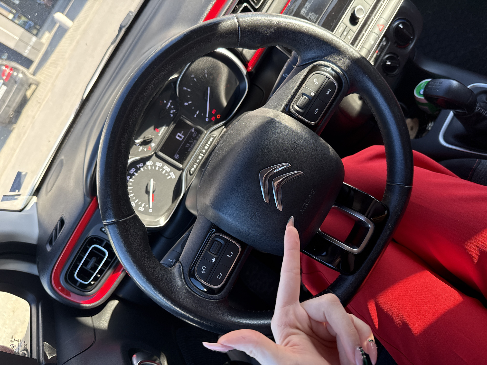 The image shows the interior of a Citroen car. The steering wheel, dashboard, and part of the center console are visible. A person's hand is pointing towards the Citroen logo on the steering wheel. The person is wearing red clothing. The instrument panel is visible behind the steering wheel. Based on context clues found in previous turns, the location is assumed to be Plovdiv, Bulgaria.