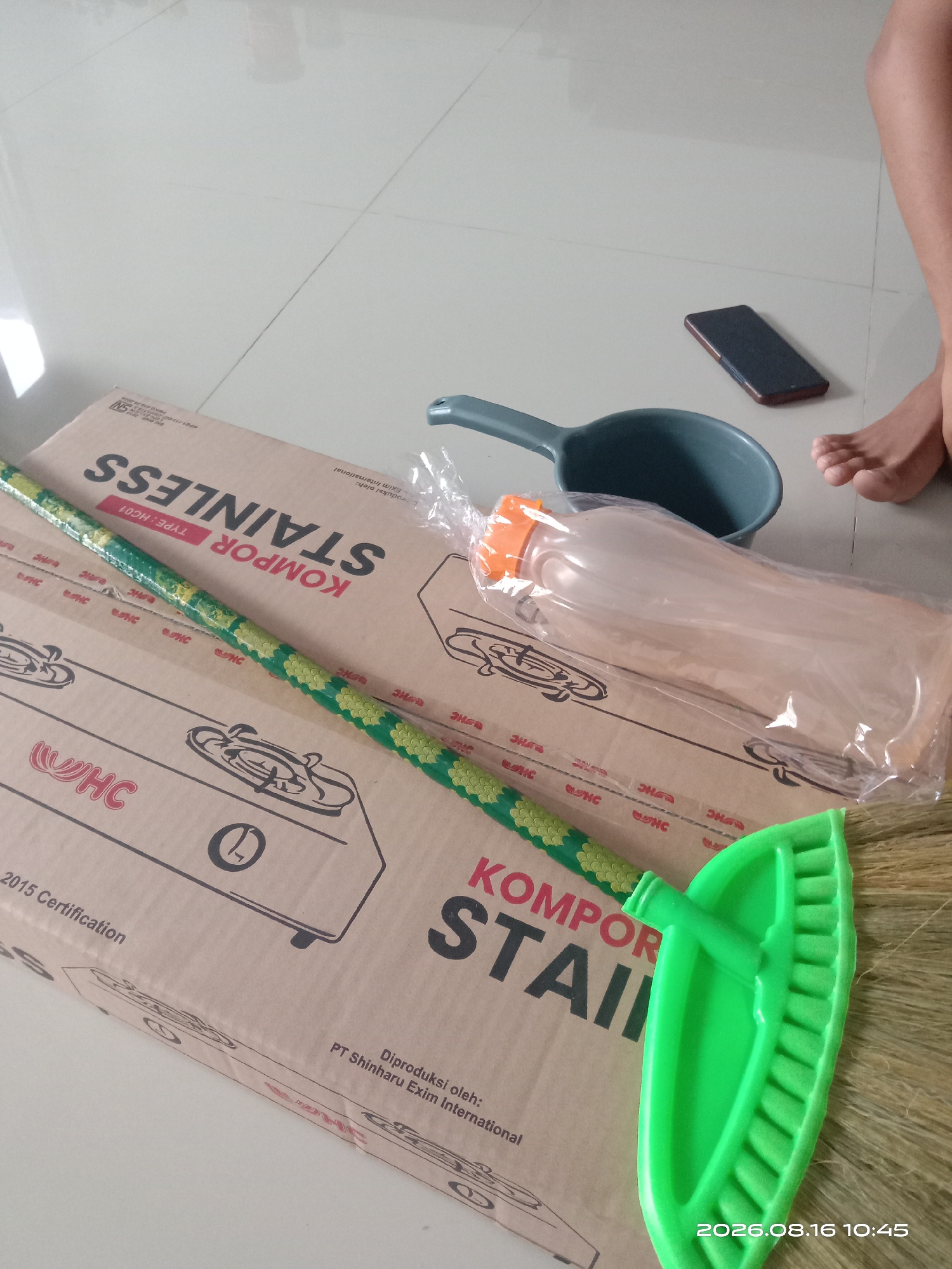 The image depicts a scene on a tiled floor, possibly in Banyuarang, Indonesia. A large cardboard box, labeled "KOMPOR STAINLESS," is prominently displayed, suggesting it contains a stove. To the left of the box is a green broom with yellow accents, lying horizontally across it. On the right side of the box, a blue ladle is partially visible, next to a clear plastic-wrapped object that appears to be a bottle with an orange cap. A dark-colored smartphone rests on the floor near the ladle and a person's bare feet, indicating a casual setting. The lighting suggests it is daytime. The overall scene appears to be an indoor domestic or commercial space where cleaning supplies and kitchen equipment are present.