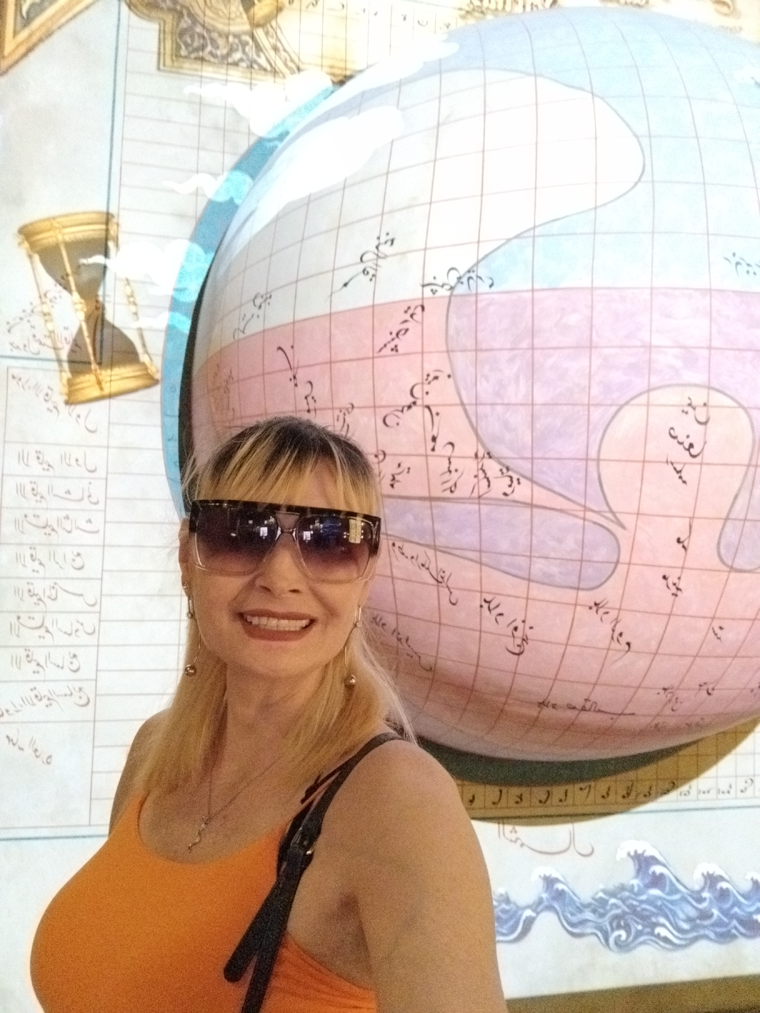 A woman with blonde hair and large sunglasses smiles at the camera, standing in front of a large, stylized globe with a map on it. The globe is depicted in pink and blue, with a grid pattern superimposed on it. There are Arabic inscriptions on the globe and on the wall behind it, which also features an hourglass illustration. The woman is wearing an orange tank top and a necklace. A dark strap of a bag is visible over her shoulder. The overall scene appears to be in a museum or exhibition space.
