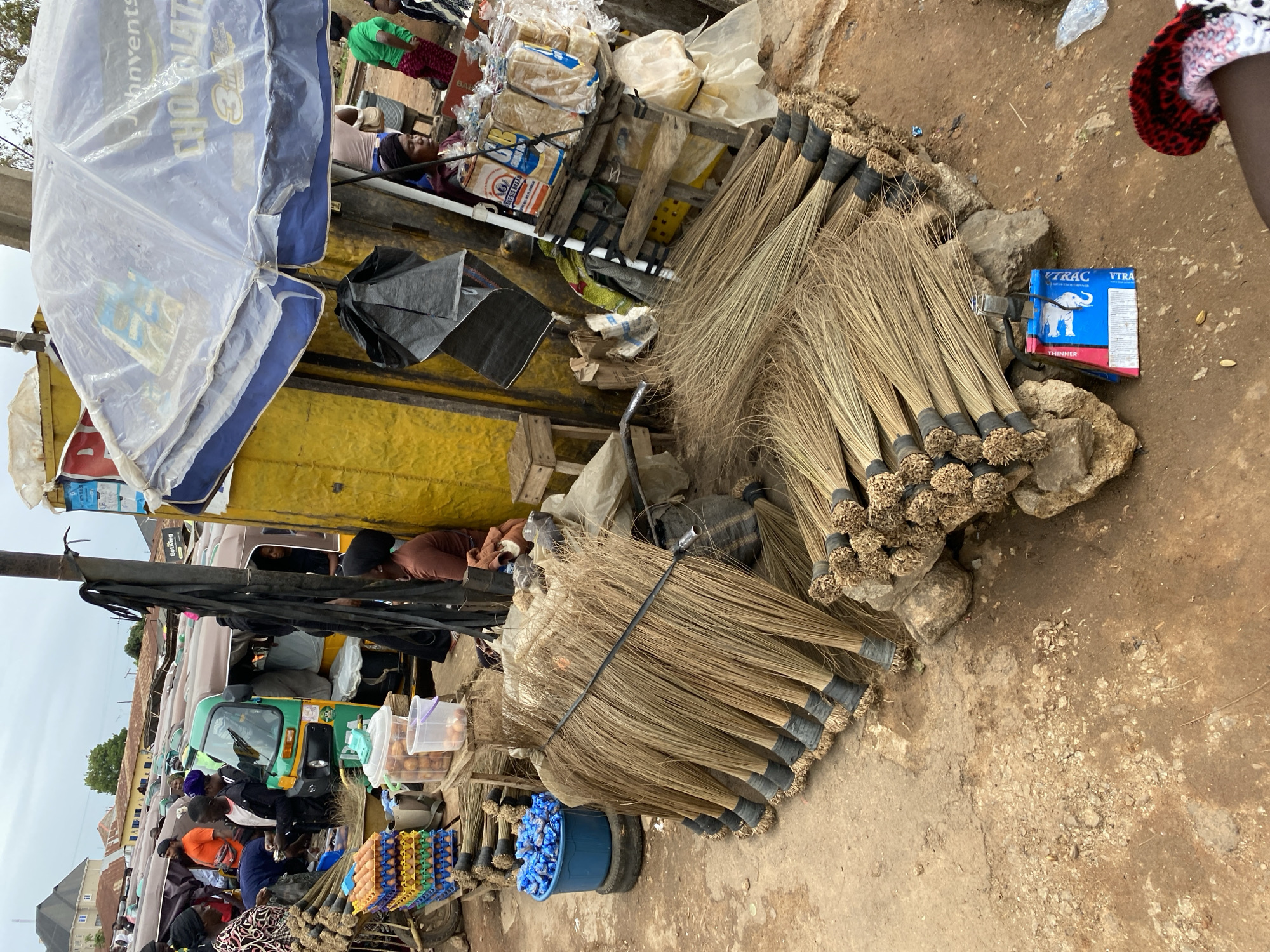 The image depicts an outdoor market scene in Bukuru, Nigeria. In the foreground, numerous brooms made of dried grass with black bindings are displayed, some stacked on top of each other, others arranged in bundles. To the left, a blue bucket filled with plastic bags is visible, along with stacks of egg cartons. Behind these, several auto-rickshaws are parked in a row. A yellow structure dominates the middle ground, with a blue and white umbrella covering a stall. Several individuals are present, some appearing to be vendors and customers. The ground is dirt and rough concrete, indicative of an unpaved market area.