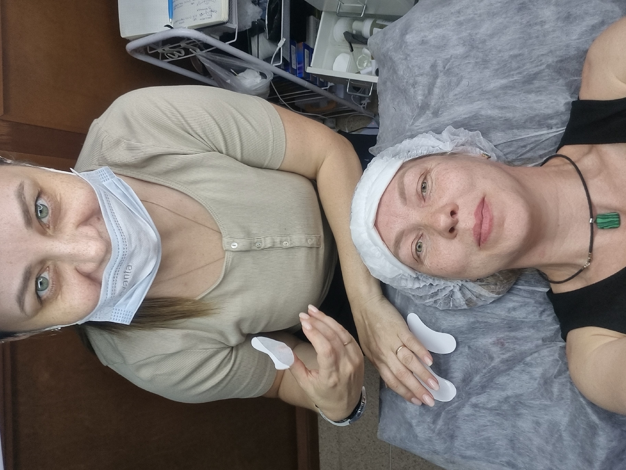 A medium, overhead shot shows two Caucasian women in a room with medical equipment. One woman is lying down with a white disposable hairnet on her head. She has light skin, light eyes, and freckles. She is wearing a black tank top and a black cord necklace with a green and black rectangular pendant. She is lying on a crinkled gray cloth. The second woman is standing and holding two white, tear-shaped objects, one in each hand. She is wearing a beige button-down shirt, a white surgical mask, and a watch with a white and black band. The background includes a light-colored wood cabinet, a metal shelf with medical supplies, and notebooks. The location is Chaikovsky, Russia.