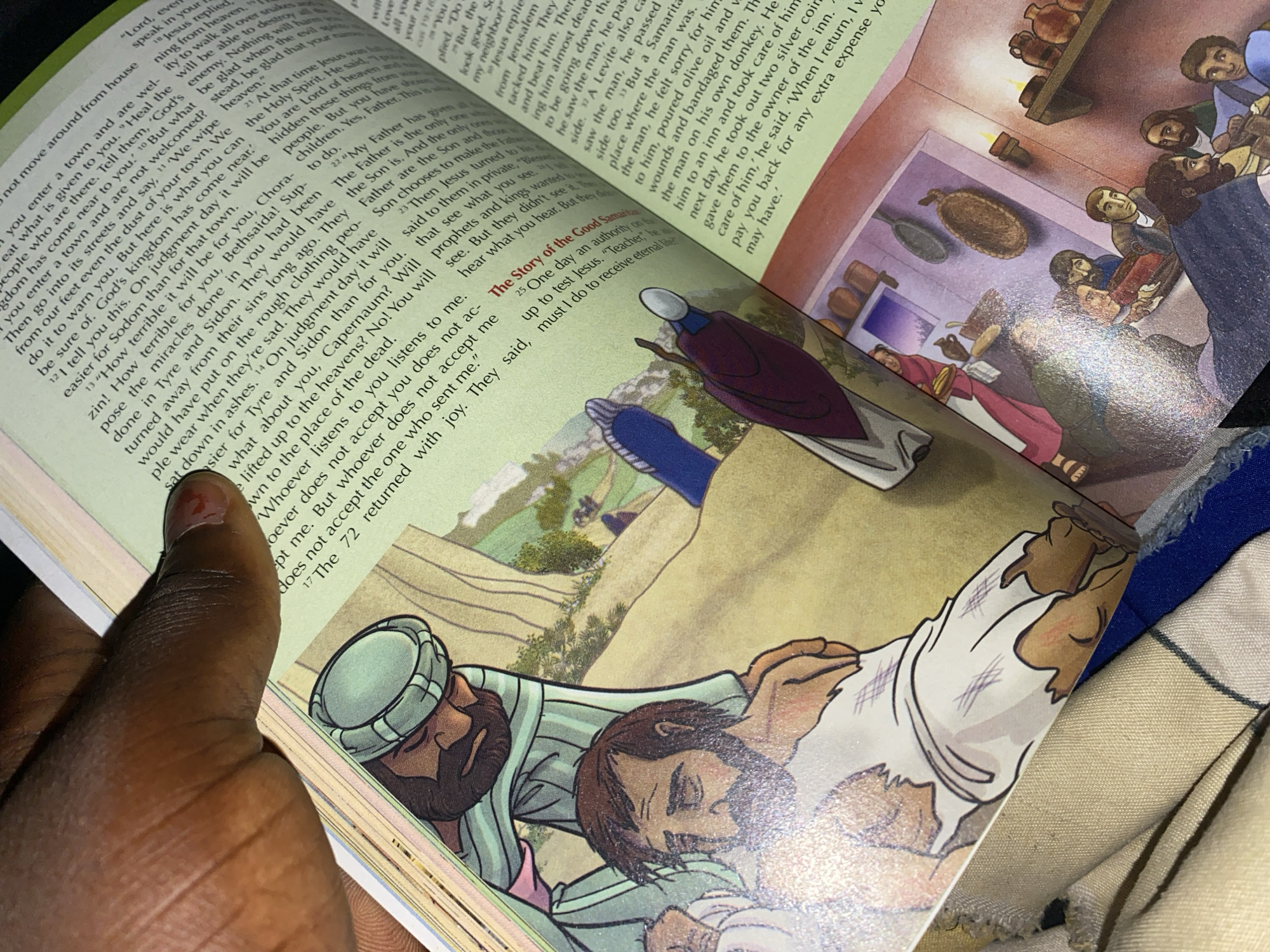 The image is a close-up shot of an open book, likely a Bible, with illustrations. The book is angled, with a person's thumb and forefinger, displaying a dark skin tone and red nail polish, holding down the top of the left page.
The illustration on the left page depicts two men. The top man is wearing a light blue garment and a light blue turban. He is leaning over and has his left hand on the shoulder of the man below him. The bottom man is lying down, wearing tattered white clothing. His head is turned to the side, and he appears to be injured or unwell. The background on this page is a light tan, with some faint green and brown elements that suggest a landscape.
The right page of the illustration shows a scene inside a building with an arched doorway. A woman in a pink garment is visible in the background, carrying a tray of food. Several men are seated around a table in the foreground, engaged in conversation or eating. The room has shelves with pottery and utensils. The text on this page is in English, with a heading "The Story of the Good Samaritan."
The surrounding environment, as visible in the background of the book, appears to be a stylized depiction of an ancient city or village, with some hills and trees. There is no specific indication of the location being Jauro Yinu, Nigeria, in the image itself.