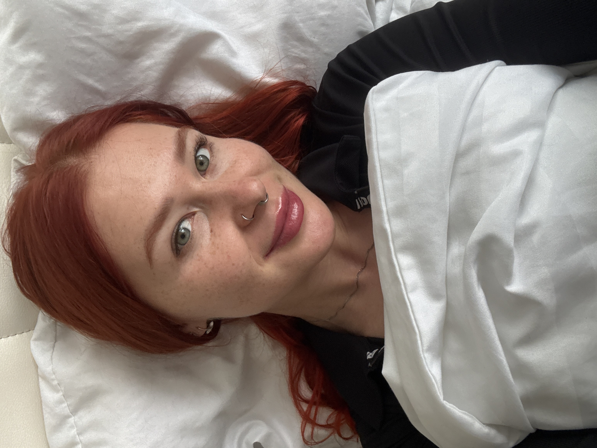 A person with red hair, fair skin, and freckles is lying on a white sheet. The person is wearing a black shirt and has a silver nose ring. A white pillow is partially covering the person's body. The location is not identifiable as Rostov-on-Don, Russia from the image.