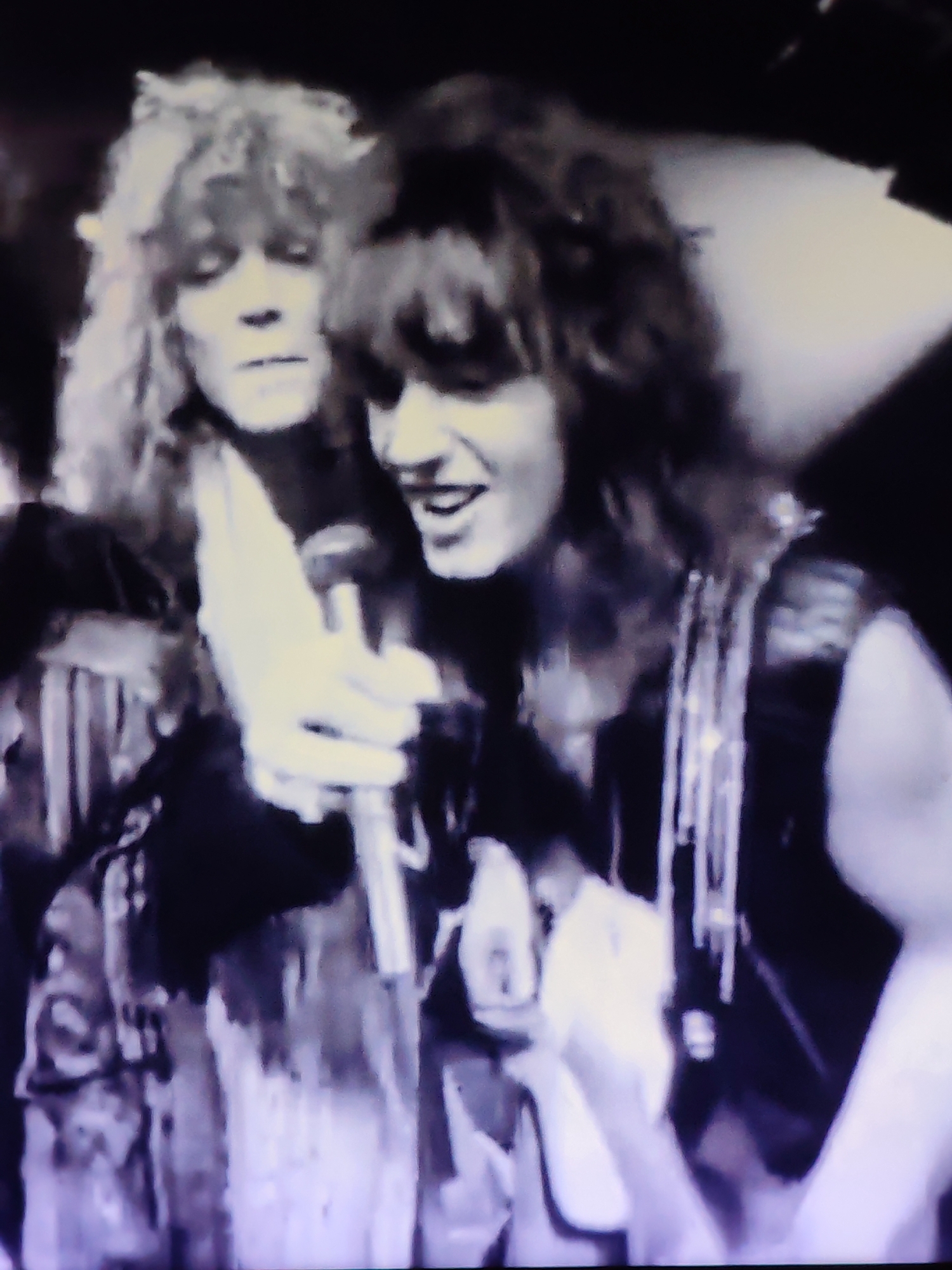 The image shows two individuals in what appears to be a performance setting. The person on the left has long, wavy blonde hair and is looking down, with their face partially obscured. The person on the right has darker, wavy hair and is looking towards the viewer, smiling and holding a microphone. Both individuals are wearing clothing that suggests a rock music performance, with decorative elements on the garments. The background is dark and out of focus, with some vertical lines visible that could be stage lighting or curtains. The image quality is low, with a grainy appearance typical of older video recordings. There is no indication of the city of Vladimir, Russia, or any specific location within it.