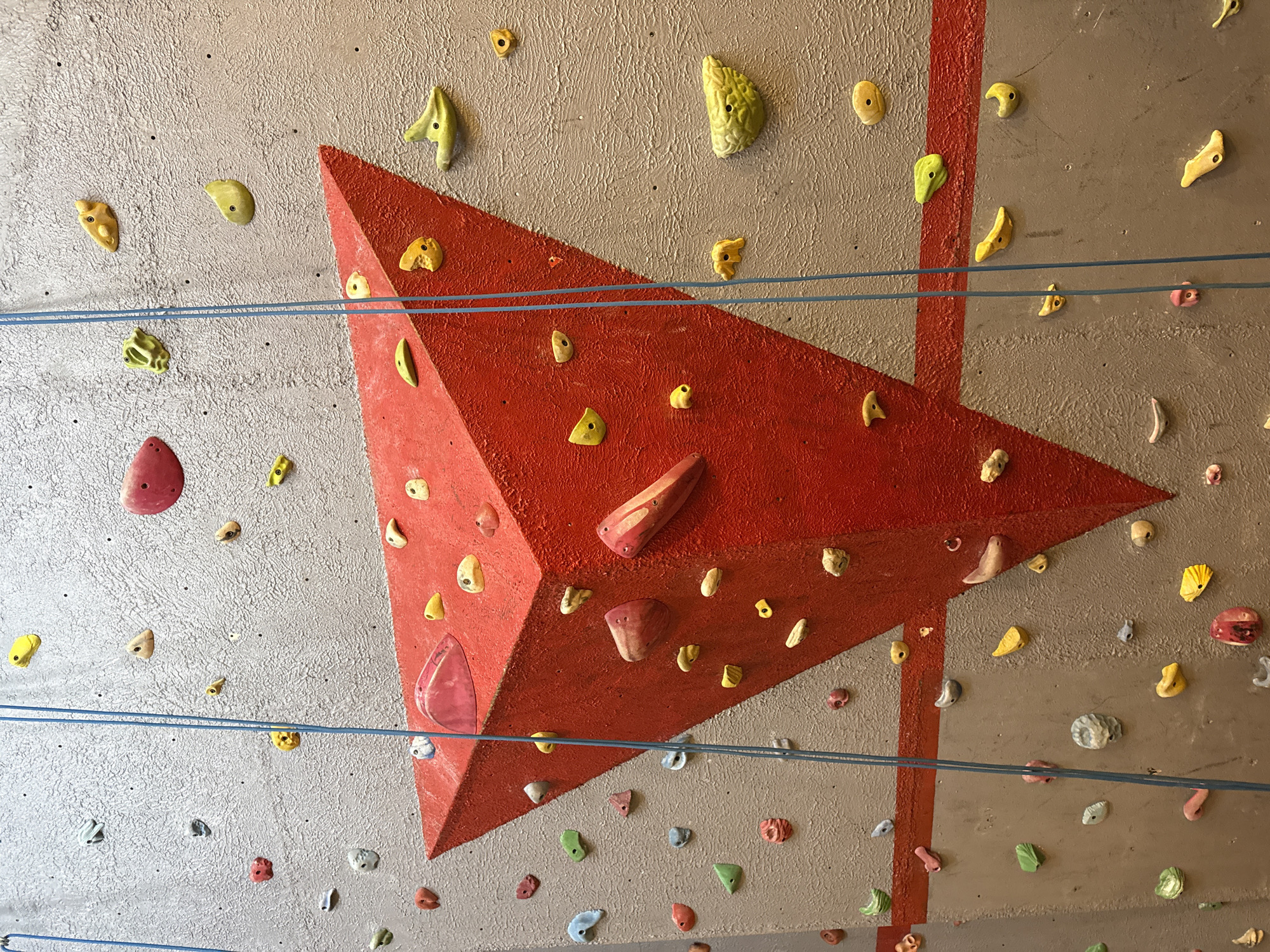 The image shows a climbing wall with various holds attached. The wall appears to be made of a textured, grey-colored material. A large, red, triangular prism is attached to the wall. Climbing holds of various shapes, sizes, and colors (yellow, pink, white, blue, green) are affixed to both the wall and the red prism. Two horizontal blue ropes extend across the wall. A red vertical stripe also runs along the wall, intersecting with the ropes. The climbing wall is located at the Volcanic Park in Puebla, Mexico.
