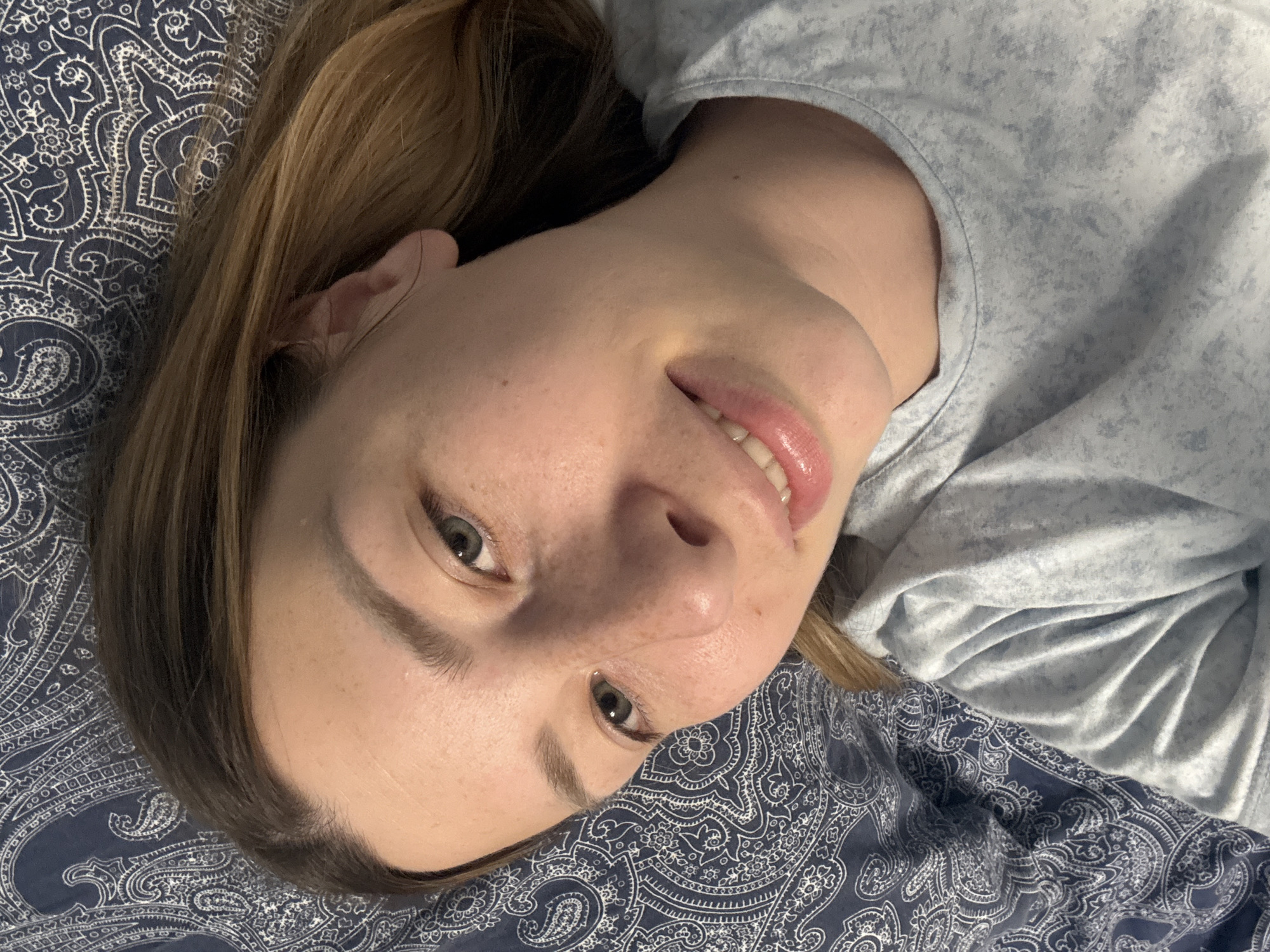 A young woman with fair skin and light brown hair is lying on a patterned blue and white bedspread, possibly in her bedroom. She is wearing a light blue shirt. The focus is on her face, which is tilted towards the camera, with a slight smile on her lips. Her eyes are open and looking directly at the viewer. The lighting is soft and diffused. There is no indication of the location being Moscow, Russia.