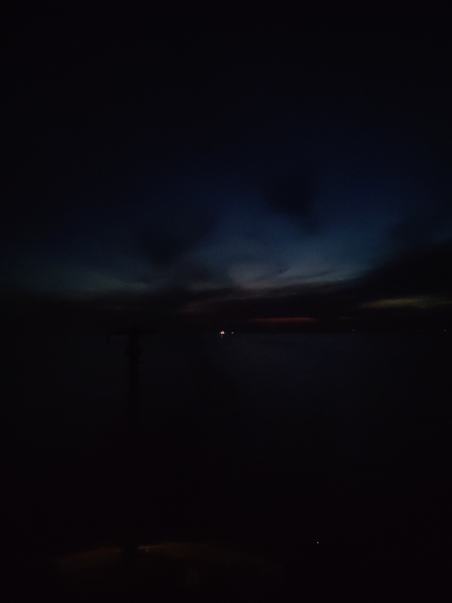The image depicts a dark, likely nighttime, scene in Bol'shie Mkhi, Russia. The sky is a gradient of dark blues and blacks, suggesting a late hour or overcast conditions. A body of water, possibly a lake or river, stretches across the midground, appearing almost entirely black. Faint lights are visible on the horizon, potentially from a distant city or boats. In the foreground, darker silhouettes imply the presence of objects like a pole.