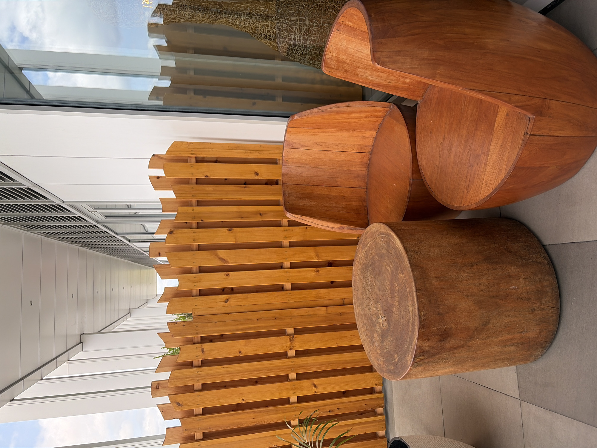 The image depicts an outdoor seating area with a wooden fence, two wooden chairs, and a wooden stool. The chairs are curved and modern in design, while the stool is a solid cylindrical log. The wooden fence is made of vertical planks and has a natural finish. The setting appears to be a balcony or patio with a tiled floor and a glass wall in the background. The lighting suggests it is daytime. The scene is devoid of people or any obvious activity. The overall impression is one of a serene and naturalistic outdoor space. The presence of the fence and the reflective glass wall suggest an urban or semi-urban environment, possibly a hotel or residential complex. While the provided information indicates Malé, Maldives, there are no specific visual cues in the image to confirm this location.