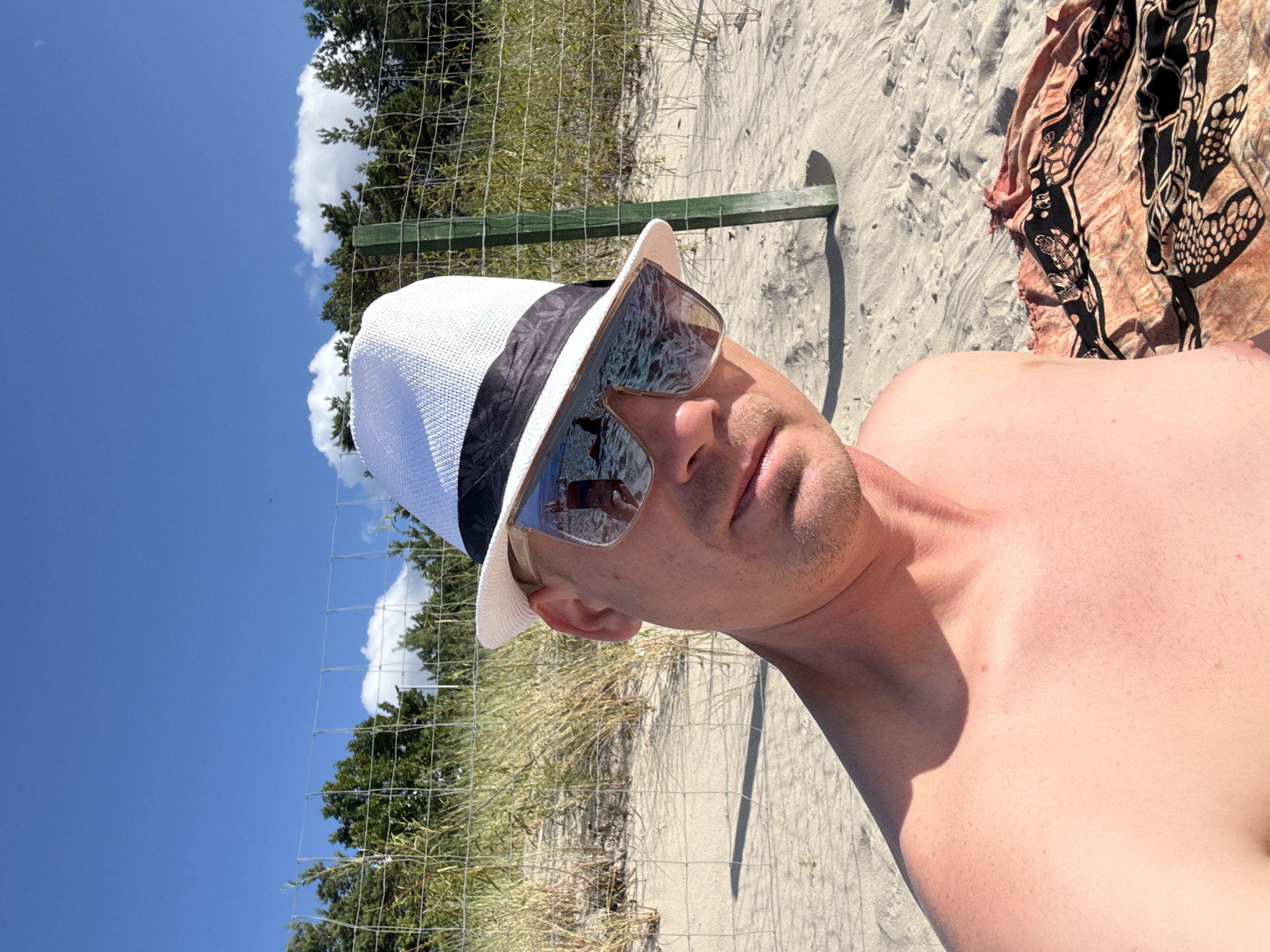 This is a first-person perspective selfie taken at a beach in Międzyzdroje, Poland. The subject is a fair-skinned man wearing a white fedora with a black band and large, square-shaped sunglasses. The sunglasses reflect the beach scene, showing a body of water and a person in the water. The man is shirtless, with his bare shoulder and upper chest visible. He is sitting on a sandy beach, with a colorful towel or sarong with a turtle print partially visible in the lower right corner. Behind him, a wire fence separates the beach from a line of trees and vegetation. The sky is blue with scattered white clouds. A green wooden post is visible near the fence. The lighting suggests it is daytime, with bright sunlight casting shadows on the sand.
