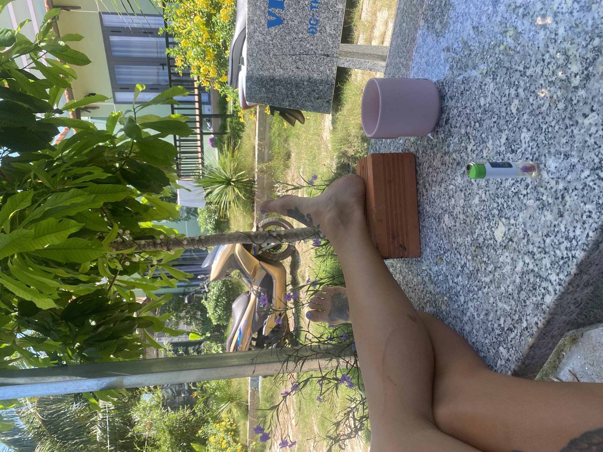 In Mũi Né, Vietnam, on a sunny day, a person's legs and foot are crossed in the foreground, resting on a stone table. The foot has a tattoo on the sole and is propped on a brick. Nearby, a pink mug and a lighter are visible on the table. In the background, a golden scooter is parked on a grassy area with purple flowers and lush green trees. A light green building with a balcony and railings is visible behind the scooter. The overall scene suggests a relaxed outdoor setting, possibly a resort or guesthouse in a tropical location.
