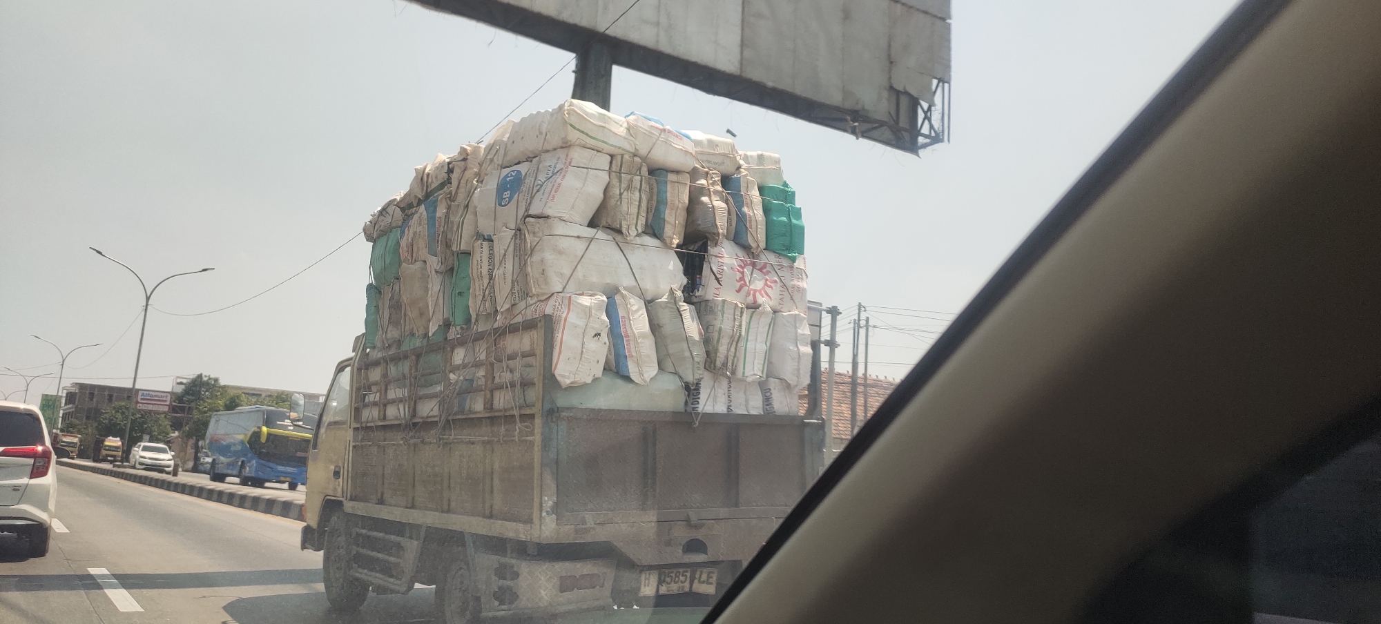 This is a medium-shot, eye-level photograph taken from a vehicle, showing a busy street scene in Sedo Lor, Indonesia. The primary subject is a heavily overloaded truck carrying a towering stack of sacks, likely agricultural produce or raw materials, secured with ropes. The sacks are of various colors, predominantly white and light blue, with some green ones interspersed. The truck is a light-colored flatbed with a metal railing system. The license plate is partially visible and reads "W 9585 LE."
The truck is in motion, traveling on a paved road with lane markings. Several other vehicles are visible on the road, including a white car in the foreground on the left, a blue and yellow bus to the left of the truck, and other cars further down the road. In the background, there are buildings, including what appears to be a storefront with an "Alfamart" sign, trees, and utility poles with overhead wires. The sky is overcast, suggesting a bright, possibly hazy day. The overall impression is a snapshot of daily life and traffic in an Indonesian town.
