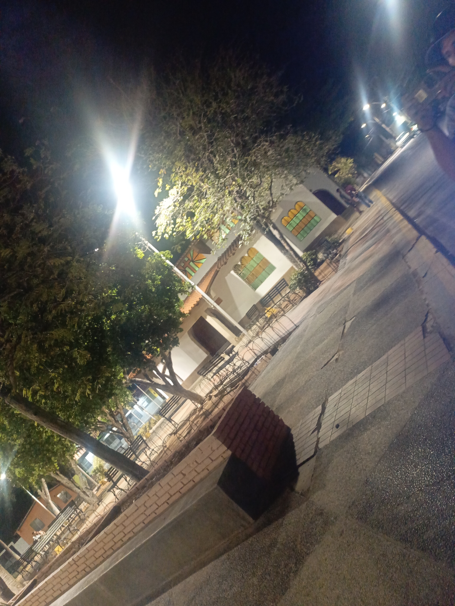 The image depicts a nighttime scene in Altagracia, Venezuela. A white building with arched windows featuring orange, green, and yellow glass panes is visible. A tree with dense foliage partially obscures the building. A brick structure is present in the foreground. The ground is paved with a combination of concrete and tile. Benches are lined up along the building. A bright street light illuminates the scene. Figures are seen walking in the distance.
