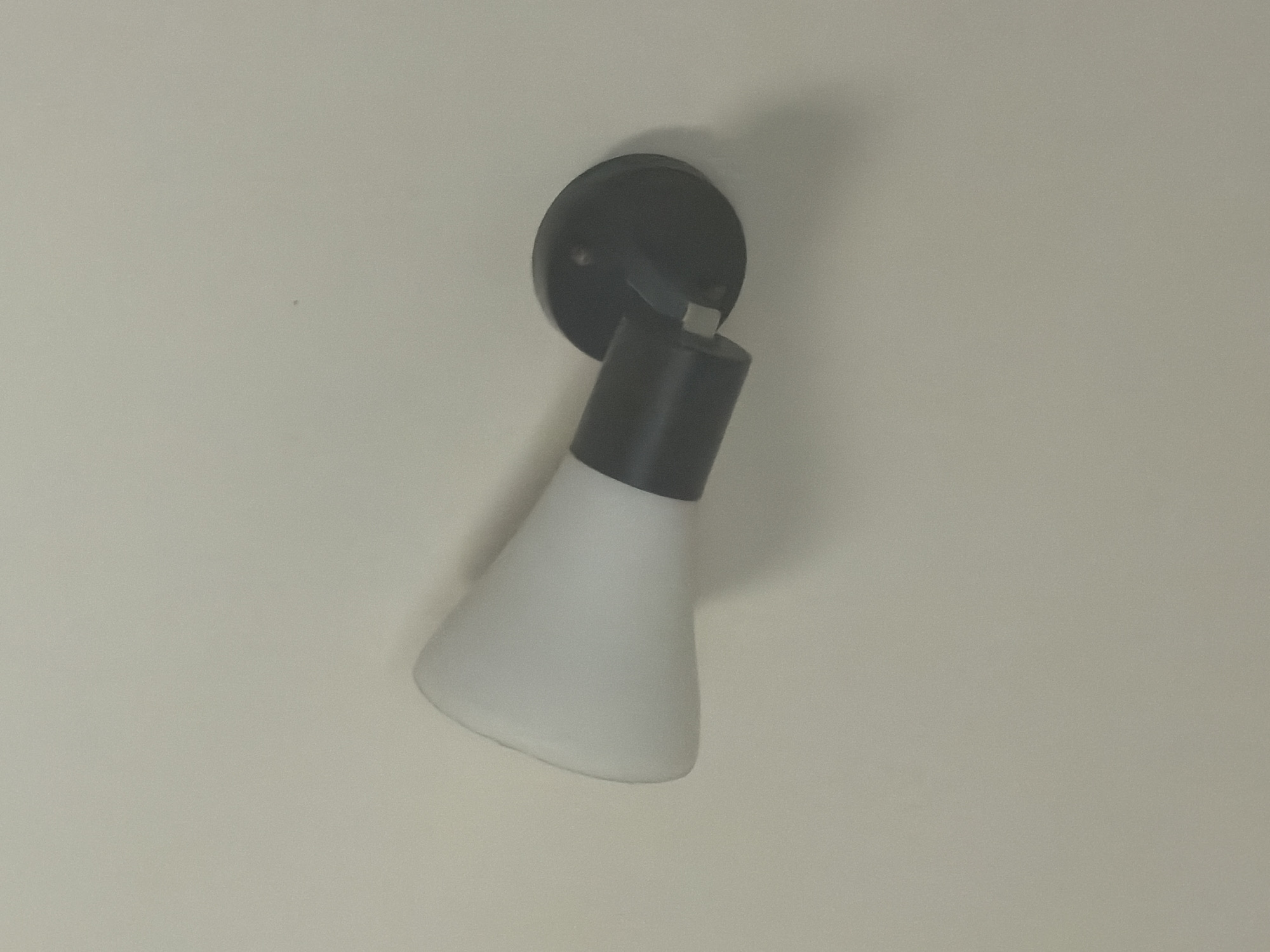 The image shows a close-up view of a black and white wall-mounted light fixture against a neutral-colored wall. The fixture consists of a circular black base attached to the wall, from which a black cylindrical arm extends. Attached to the arm is a white, conical lampshade. The lighting appears to be soft, suggesting it might be indoors or during a time of day with diffused natural light. The overall impression is that of a functional and modern interior design element. The context of Yerevan, Armenia, provides a geographical reference, but there are no specific visual cues within the image to indicate this location. No people are visible, and no text is present.