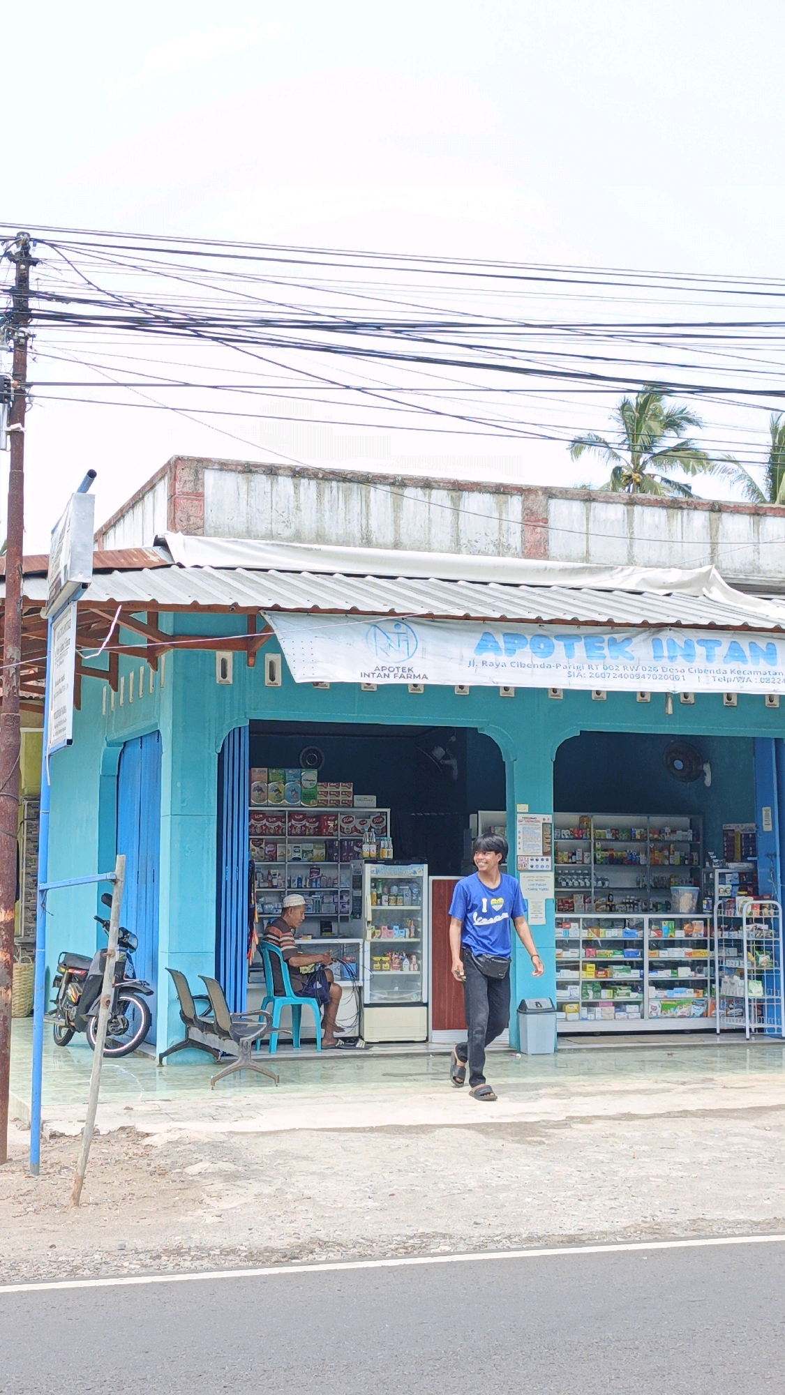 This image depicts a street view in Cibenda, Indonesia, likely in front of a pharmacy. The pharmacy, painted a bright turquoise, has a white awning with text that reads "APOTEK INTAN FARMA" and includes an address: "Jl. Raya Cibenda-Parigi RT 002 RW 026 Desa Cibenda Kecamatan Cibodas Kab. Tangerang, Banten". A motorcycle is parked to the left of the pharmacy.
Inside the pharmacy, shelves are stocked with various products, and a man is seated on a blue chair, appearing to be an employee or customer. To the right, a young man in a blue t-shirt with a "I <3 Cups" graphic and black pants walks past the pharmacy. He is wearing a black fanny pack. The street in front of the pharmacy is paved, and overhead power lines are prominent in the sky. The overall environment suggests a typical Indonesian urban or suburban setting.