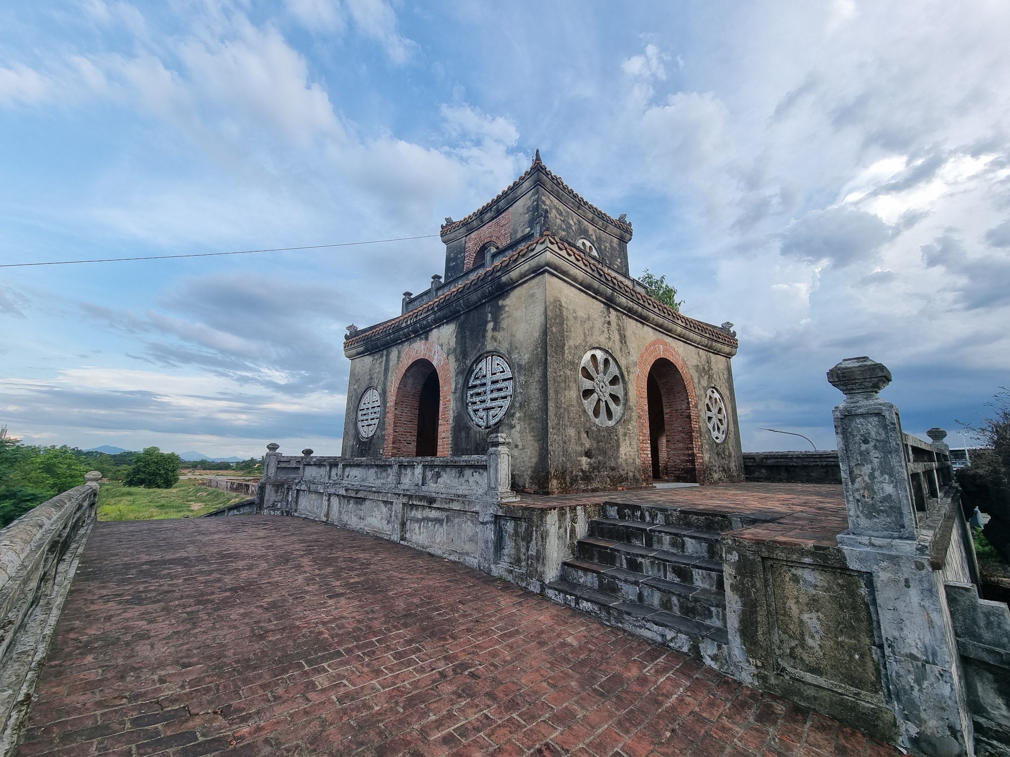 An exterior, eye-level, wide-angle shot captures an ancient, weathered, octagonal tower with a multi-tiered, tiled roof under a cloudy sky. The tower is constructed of stone and brick, with arched openings revealing dark interiors. Circular decorative motifs, possibly windows or carvings, adorn the facade. A wide brick pathway leads up to the tower, bordered by a stone balustrade. Stairs ascend from the pathway to the tower's elevated platform. Lush green foliage and distant mountains are visible in the background. The scene is devoid of human presence. This image was taken at the Phu Xuan Ward, Vietnam.