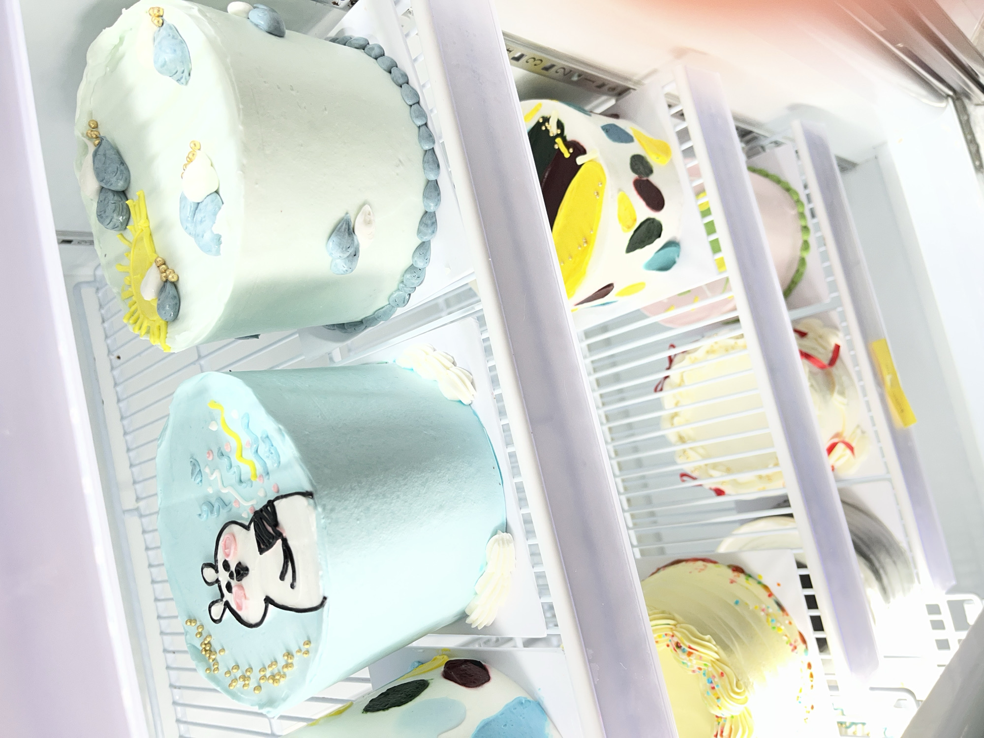 The image depicts a close-up, interior view of a refrigerator showcasing an assortment of decorative cakes arranged on wire shelves. The primary focus is on the upper shelf, which holds two distinct cakes. The cake on the left is a cylindrical, light blue frosted cake adorned with a cartoonish white character, possibly a hamster, holding a black object. Gold sprinkles and blue and pink wavy frosting details are also visible on its top. To its right sits another cylindrical cake, frosted in a pale green hue. This cake is decorated with a yellow sun motif, grey and white cloud-like fondant pieces, and gold edible beads.
Below these two cakes, the next shelf contains more cakes. One on the left is partially visible, white frosted with yellow frosting swirls and colorful sprinkles. Another cake to its right is mostly white with red ribbon accents. A third cake on this shelf is white with abstract colorful splashes of yellow, dark brown, and teal.
The lowest visible shelf shows a pink frosted cake with a green frosting border. The refrigerator's interior, characterized by white wire shelving and fluorescent lighting, is clearly visible. The context suggests this is a bakery or a display case, likely within Lhokseumawe, Indonesia, given the specified location. No people are present in the image.