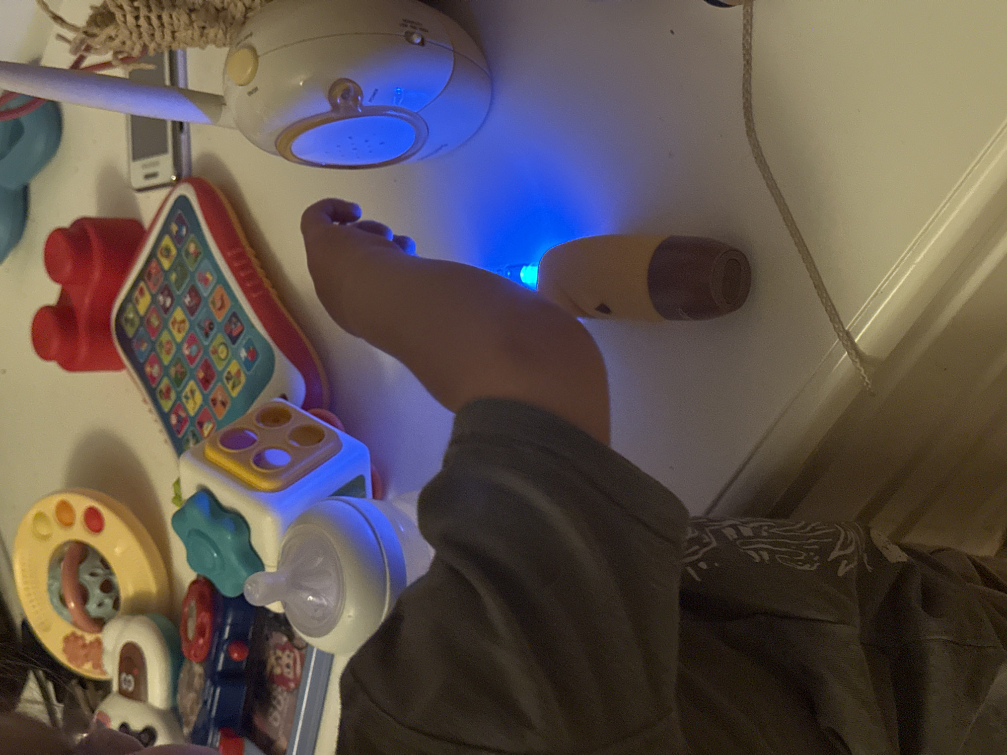 The image depicts a close-up view of a child's hands interacting with various toys on a white surface, likely a table or dresser. Several toys are visible, including a baby bottle with a blue light emanating from its base, a colorful electronic learning toy with buttons, a yellow ring-shaped toy, and red building blocks. A white baby monitor unit with a yellow button is also present. The child is wearing a dark t-shirt with a white graphic design on the sleeve. The lighting in the scene is dim, with the primary light source appearing to be the blue glow from the bottle. The image does not contain any identifiable landmarks or context that would allow for its location to be determined in Sofia, Bulgaria, or any other city.
