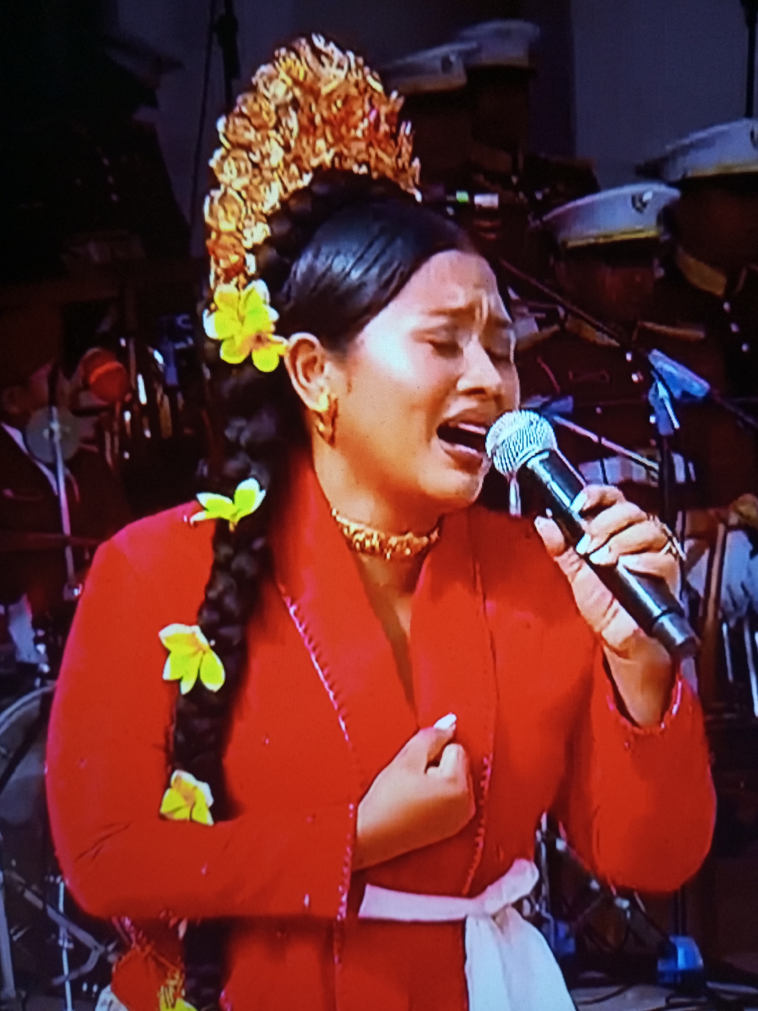 A medium shot captures a female singer performing on a stage in Banyudono, Indonesia. She is adorned in a vibrant red, traditional Indonesian outfit, complete with a white sash tied around her waist. Her dark hair is styled in a large braid, embellished with yellow flowers and an ornate golden headpiece. The singer holds a microphone to her mouth, her eyes closed and her mouth open in song. Behind her, a band is visible, with instruments and stage equipment dimly lit in the background. The overall atmosphere suggests a live musical performance.