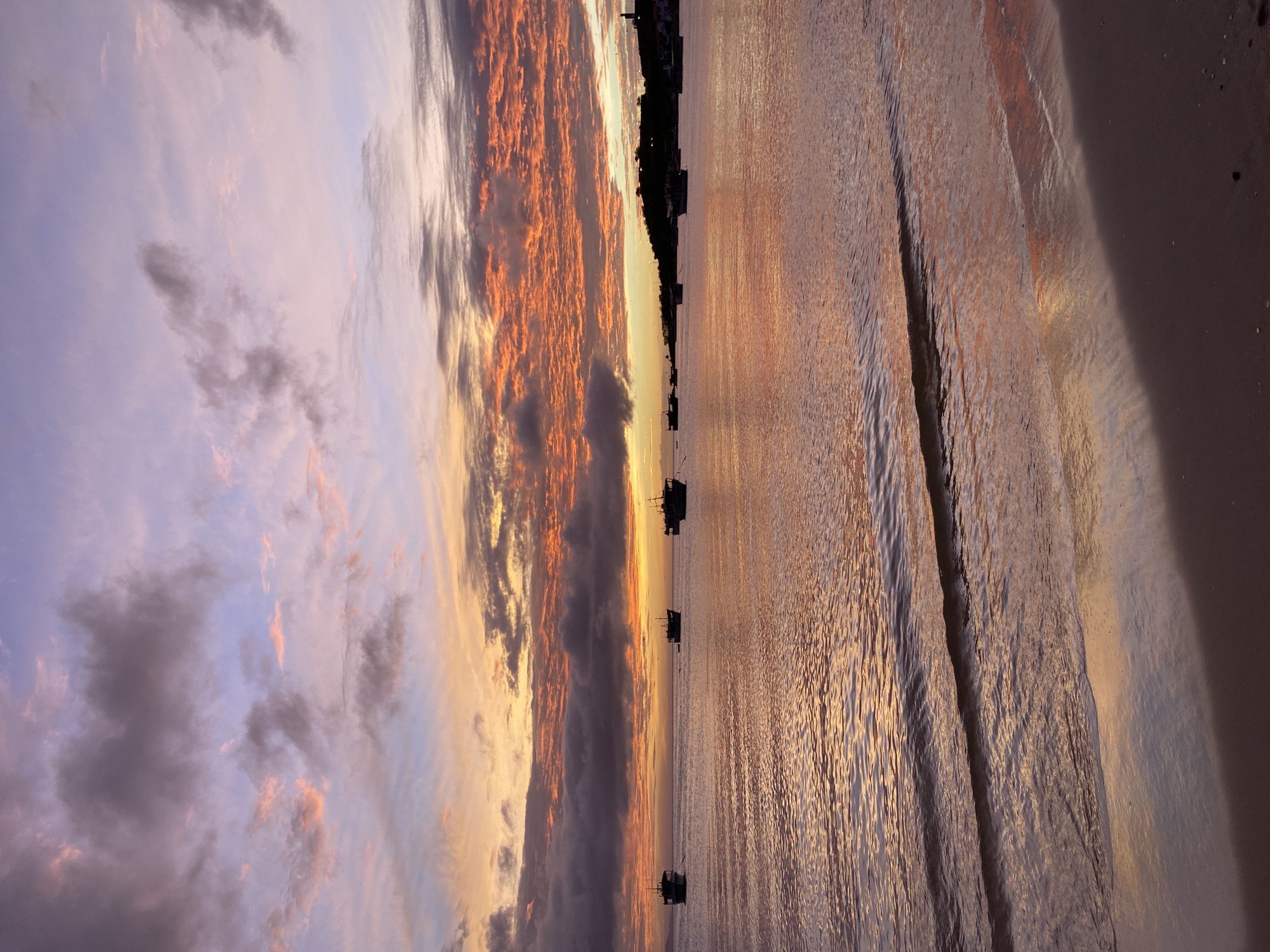 This vertical, high-angle, full-shot photograph depicts a serene sunset over the ocean in Juan Griego, Venezuela. The sky is filled with dramatic clouds, painted in hues of purple, pink, and orange, with the sun setting near the horizon. A gentle, rippling ocean stretches towards the background, reflecting the warm colors of the sky. Several boats are anchored in the water, silhouetted against the colorful twilight. In the far distance, a dimly lit coastline is visible, with a few buildings and structures suggesting a populated area. The foreground shows a sandy beach with the edge of the water gently lapping at the shore. The overall mood is peaceful and tranquil, capturing the beauty of a coastal sunset.