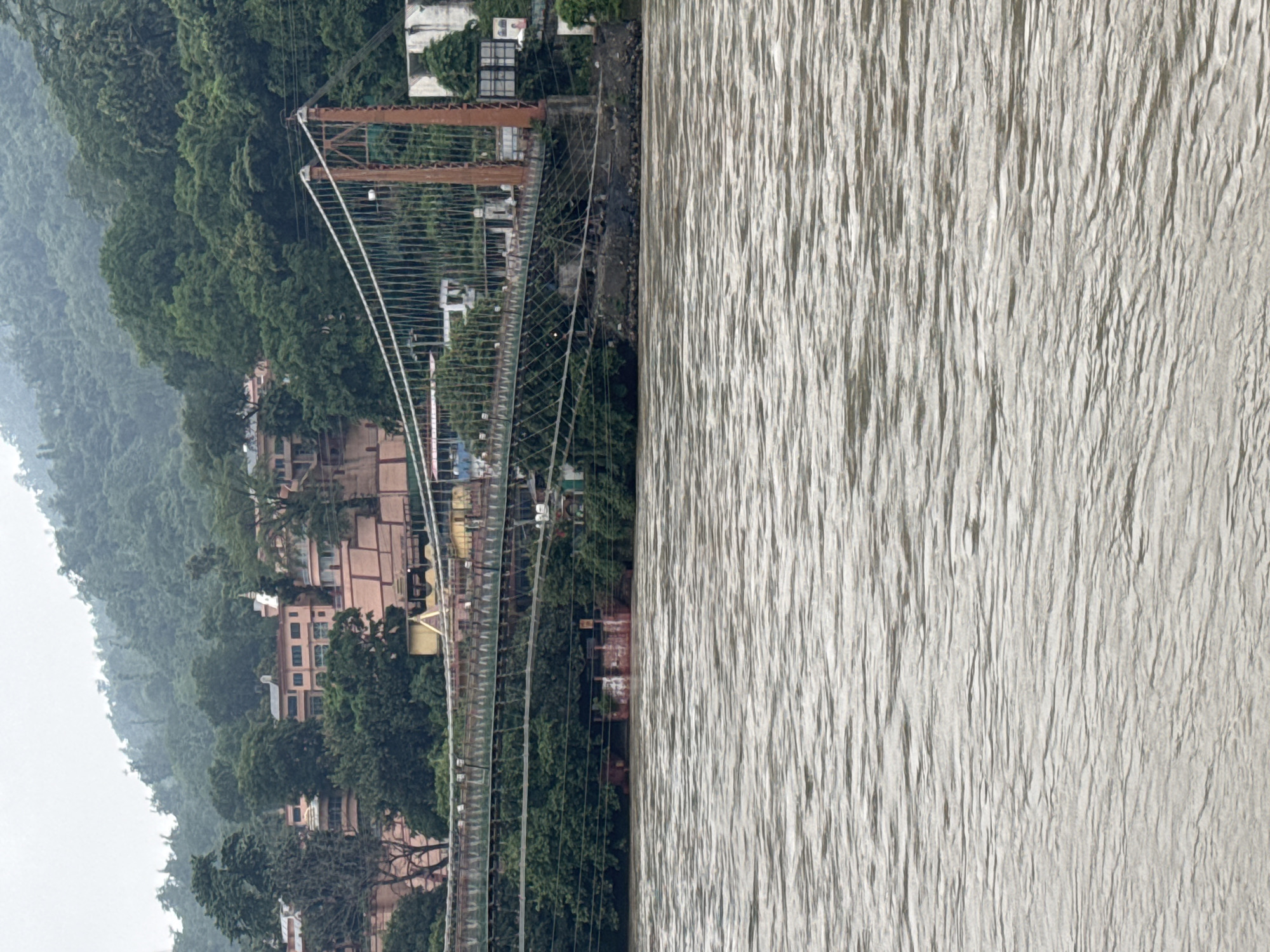 The image shows a suspension bridge crossing a wide, brown river. The bridge has a metal superstructure supported by two large, rust-colored pillars. The bridge deck appears to be made of metal grating and is lined with railings. Dense green foliage covers the riverbanks on either side of the bridge. In the background, there are several multi-story buildings with light-colored facades, possibly temples or ashrams, nestled amongst lush green trees on a hillside. The sky is overcast, contributing to a muted, natural light. The water in the foreground is textured with ripples, suggesting a moderate current. This scene is characteristic of Rishikesh, India, specifically the area near the iconic Ram Jhula or Lakshman Jhula bridges, which span the Ganges River amidst the surrounding hills and spiritual centers. There are no people or discernible actions captured in this static view.
