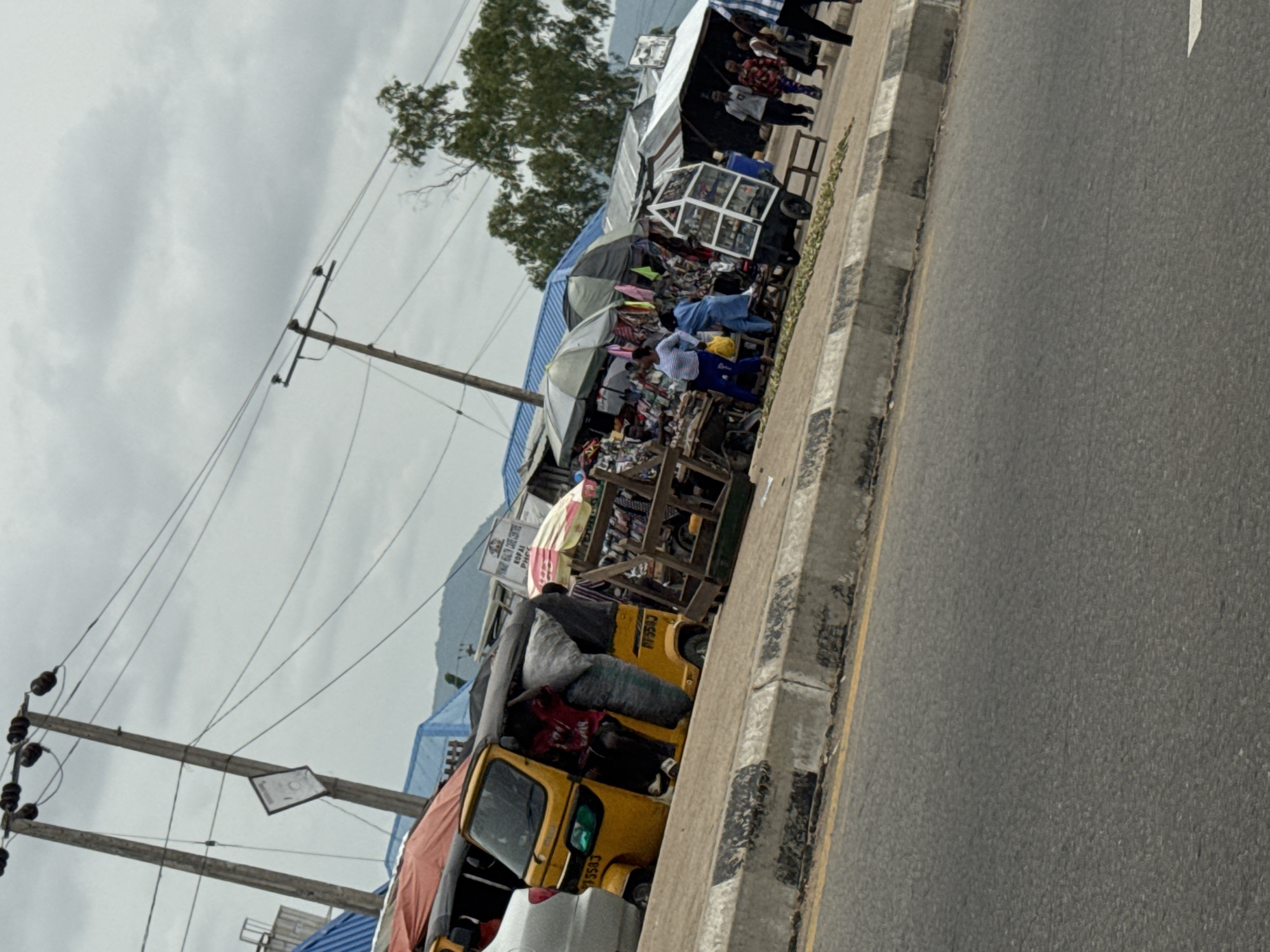 This is a street scene in Jalingo, Nigeria, with a focus on a roadside market. In the foreground, a yellow autorickshaw, also known as a "Keke Napep", is parked on the side of a paved road. The Keke appears to be loaded with goods, with a person partially visible inside, seemingly adjusting the cargo. The vehicle has the license plate "PF583".
The background reveals a bustling marketplace with numerous stalls and vendors under colorful awnings and umbrellas. People are seen browsing and interacting, suggesting a busy commercial area. Various goods are displayed, including what appear to be clothing, household items, and potentially food. A wooden cart filled with items is also visible, as is a white display rack with multiple shelves, likely showcasing smaller products.
Overhead, electrical wires are strung between utility poles, indicating the presence of power infrastructure in the area. The sky is overcast with clouds, suggesting a diffused light condition. In the distance, hills or mountains can be seen under the cloudy sky, providing a natural backdrop to the urban setting. The overall impression is that of a typical day in a vibrant, active market in Jalingo, Nigeria.