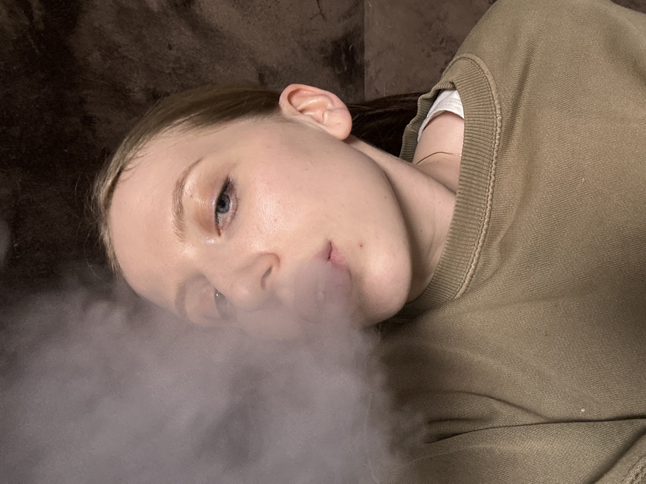 A close-up, chest-up portrait depicts a female subject exhaling a cloud of light grey vapor or smoke. The subject, positioned slightly upwards towards the camera, has light skin, light-colored eyes, and light brown or blonde hair pulled back. Her lips are slightly puckered as the vapor emanates from her mouth, obscuring the left side of her face. She is wearing a light olive or khaki-colored crewneck sweatshirt over a visible white undershirt. The background is a dark, out-of-focus, textured surface, possibly a wall or fabric, in a dark brown or black hue. The lighting illuminates the subject from the front. The specific city location of Chaikovsky, Russia, is not discernable from the visual evidence within the image.