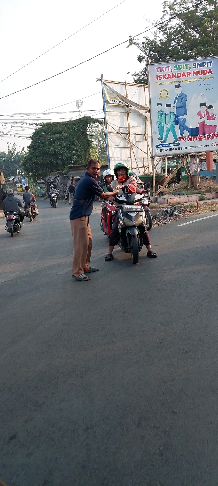 In a street scene in Satriamekar, Indonesia, two men are interacting with a motorcycle. One man, wearing a navy blue shirt and tan pants, is standing on the left and appears to be talking to or gesturing towards the other man. The second man, wearing a red and black striped shirt, a helmet, and a scarf over his face, is sitting on a red and black motorcycle. He is looking towards the standing man. Another person, wearing a white helmet, is seated behind the rider on the same motorcycle. The motorcycle has a license plate that reads "B 6277 KJD". Several other motorcycles and people are visible in the background, with some riding away from the viewer. A large billboard is prominently displayed on the right side of the image, advertising "TKIT, SDIT, SMPIT ISKANDAR MUDA" with images of students and an inscription in Indonesian. The environment suggests a sunny day with clear skies, and the road surface is asphalt. Trees and some buildings are visible in the background.
