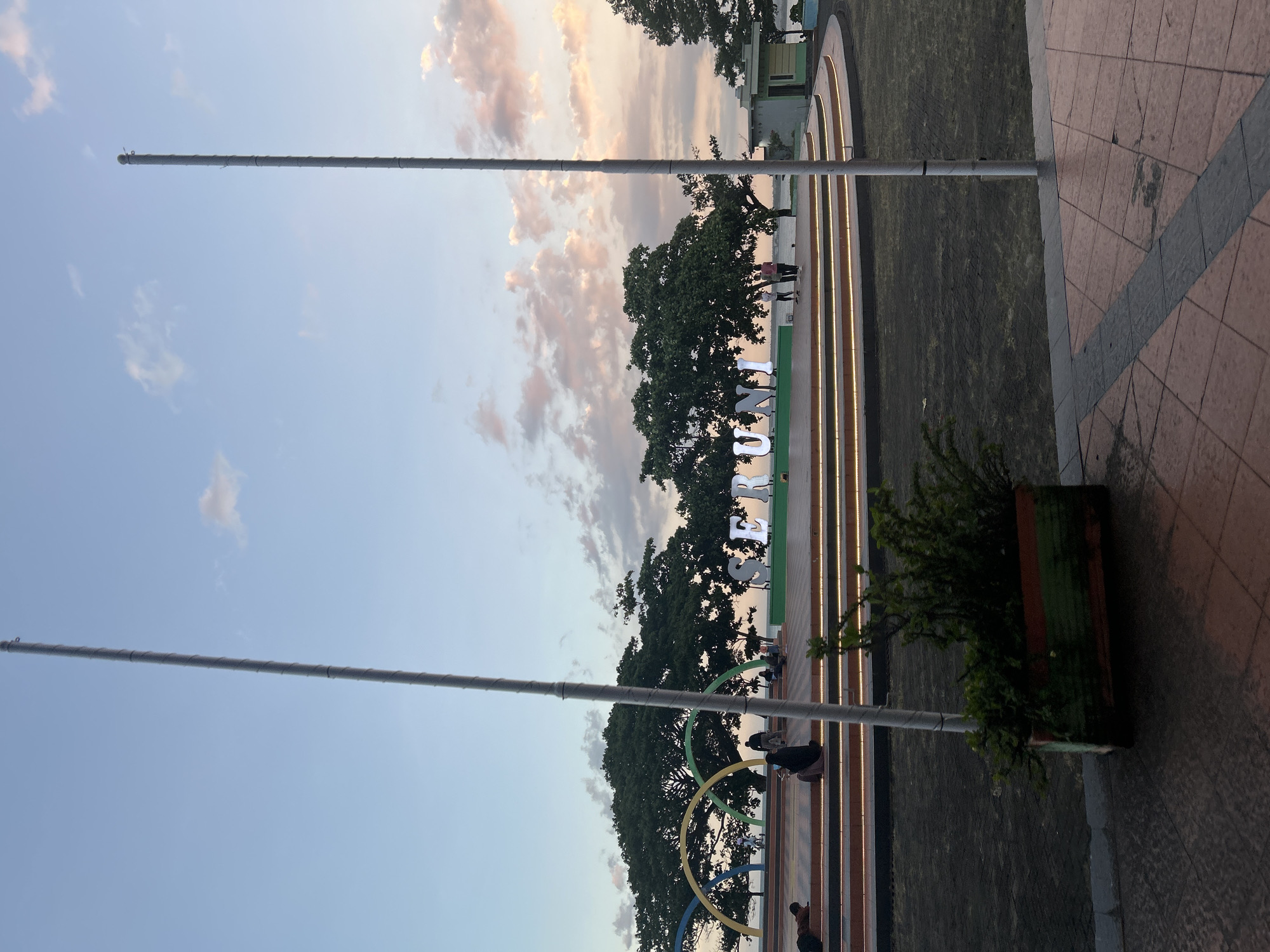 The image depicts a waterfront park in Bantaeng, Indonesia, during what appears to be late afternoon or early evening, given the soft lighting and colorful sky. In the foreground, a rectangular planter with lush green foliage sits on a tiled surface. Two tall, light-colored poles, likely flagpoles, are prominent in the scene, one on either side of the frame, with a section of their grey, textured surface visible.
The middle ground features a multi-tiered circular platform, illuminated by warm lights along its edges. Several people are present on this platform. Some are seated, while others are standing or walking. Behind the platform, large trees with dense green canopies dominate the landscape, providing shade. Across the water, which is visible beyond the trees, is a subtle hint of land or structures.
A notable feature of the park is a large, illuminated sign spelling out "SERUNI" in white, three-dimensional letters, positioned on the circular platform. To the left of the sign, a colorful arched structure, with yellow, green, and blue segments, adds a playful element to the environment. Further to the right, a small building with a light green roof and beige walls is visible. The sky above is a gradient of soft blue, with streaks of pink and white clouds, indicative of a sunset. The overall impression is a tranquil public space, likely a popular spot for recreation and relaxation in Bantaeng.