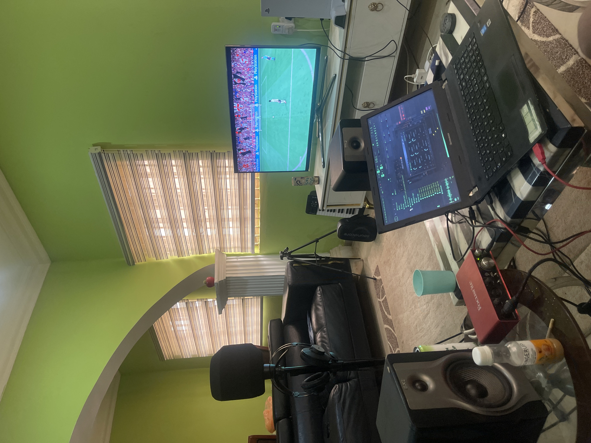The interior of a room is shown. A television is playing a soccer game with “Playstation” logos visible on the screen. A Playstation console is visible on a stand above the television. A laptop with music production software on the screen is on a table, next to the TV. Two studio monitor speakers flank the laptop. There is a red audio interface, a mint-colored cup, and a small bottle on the table, along with tangled wires. A black leather couch is in the foreground. A microphone with a pop filter is near the couch. A boom stand with a Soundcore speaker attached is behind the couch. The room is painted green and has striped blinds on the windows. An arched opening is visible on the left. The city of Abuja, Nigeria is not apparent from the image.