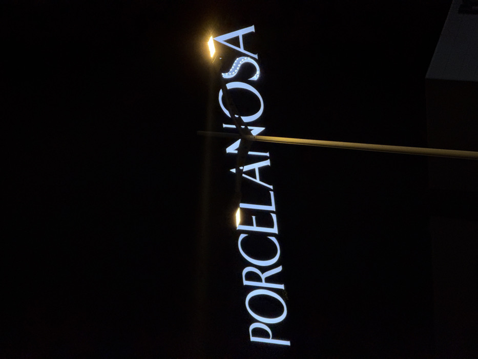 An outdoor, nighttime photograph depicts an illuminated "PORCELANOSA" sign in Polígon Industrial De Son Castello, Spain. The sign features white capital letters against a black background. A vertical, light-colored street light pole obstructs a portion of the letters "LAI" in the sign. Two active street lamps with bright, rectangular, yellow-orange light sources are visible: one above the "E" and "L" of the sign, and the other above the "O" and "S". A dark, angular building structure is discernible in the lower right corner of the frame. The sky is entirely black. No individuals or direct actions are visible.