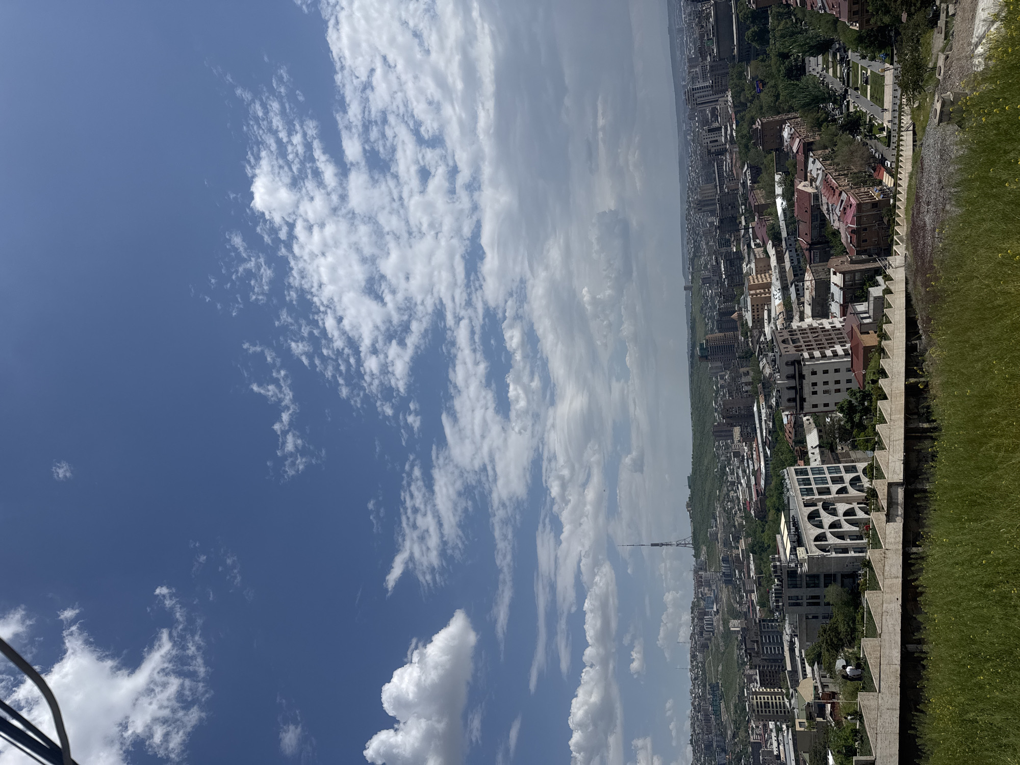 The image shows an elevated view of Yerevan, Armenia, on a sunny day. The sky is predominantly blue with numerous white clouds. In the midground, a dense urban landscape stretches into the distance, featuring a mix of buildings, trees, and roads. A telecommunications tower is visible amid the buildings. In the foreground, a retaining wall or structure with steps runs along the edge, with green grass and vegetation visible on the top.