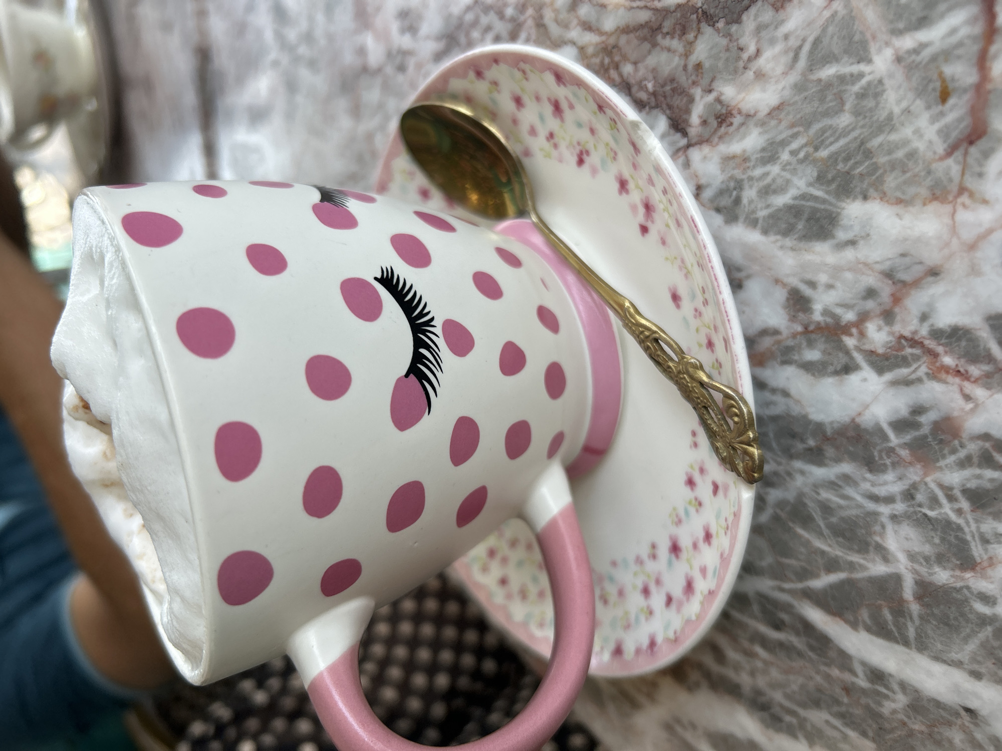 A close-up shot captures a whimsical teacup and saucer set tilted on a marbled surface. The white teacup is adorned with bright pink polka dots and a stylized black closed eye with prominent eyelashes. Its handle is a solid pink. Inside the cup, a soft, white, frothy substance, resembling whipped cream or foam, is visible. The matching saucer features a delicate pattern of small pink flowers and leaves, complemented by a pink rim. A golden spoon with an ornate handle rests on the saucer. The background is dominated by a grey and white marbled surface with reddish-brown veins. A blurred view of a person's arm and dark clothing can be seen in the distance behind the cup. There is no visible text.