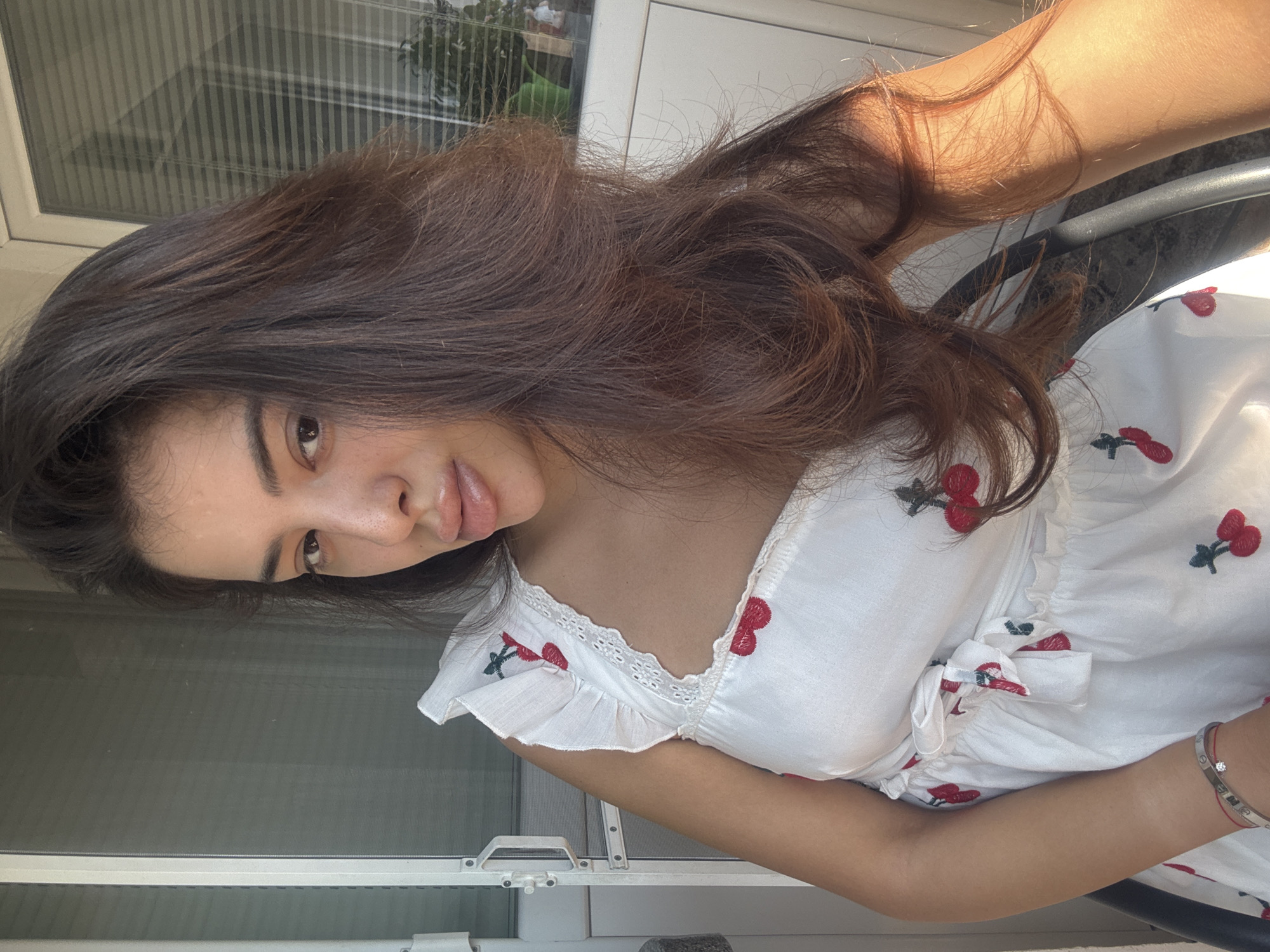 In Plovdiv, Bulgaria, a woman with long, dark brown hair is captured in a selfie. She is wearing a white dress with a cherry print. The setting appears to be outdoors, possibly on a balcony or patio, with a building and blinds visible in the background. The lighting suggests it is daytime, with sunlight illuminating the right side of her face and hair. She is looking towards the camera with a neutral expression. A bracelet adorns her left wrist. The image is taken from a medium-close shot, focusing on her upper body and face.