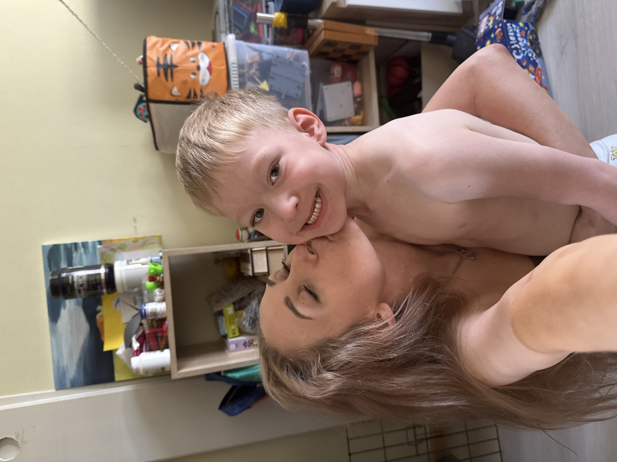 A fair-skinned woman with light brown hair is lying on her side, kissing a young, fair-skinned boy with blond hair on the cheek. The boy is smiling and looking at the camera. Both are partially unclothed. They are in an indoor setting, likely a home. In the background is a wall, shelving, and various objects including toys and storage containers. The location is not specified.