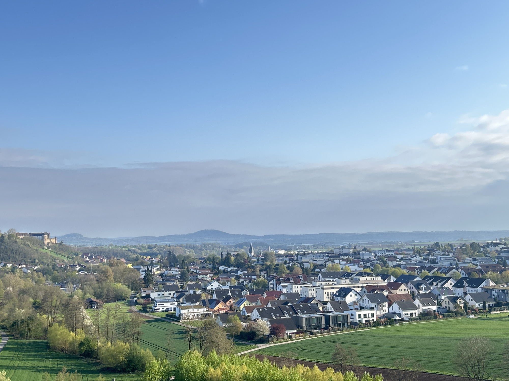 The image captures a panoramic view of a town, exhibiting a mix of residential areas and green agricultural fields. The scene is taken from an elevated perspective, providing a broad overview of the landscape. In the foreground, green fields stretch out, suggesting active agriculture. The town itself is densely packed with houses, displaying various architectural styles and colors, with visible roads weaving through the residential areas. In the background, a skyline is present with a few prominent structures like a church spire and a castle on a hill, indicating historical significance. Distant hills and a hazy sky complete the backdrop, creating a sense of depth and a serene atmosphere. The overall environment is a blend of rural and urban elements, suggesting a small to medium-sized town with a blend of historical and modern characteristics.