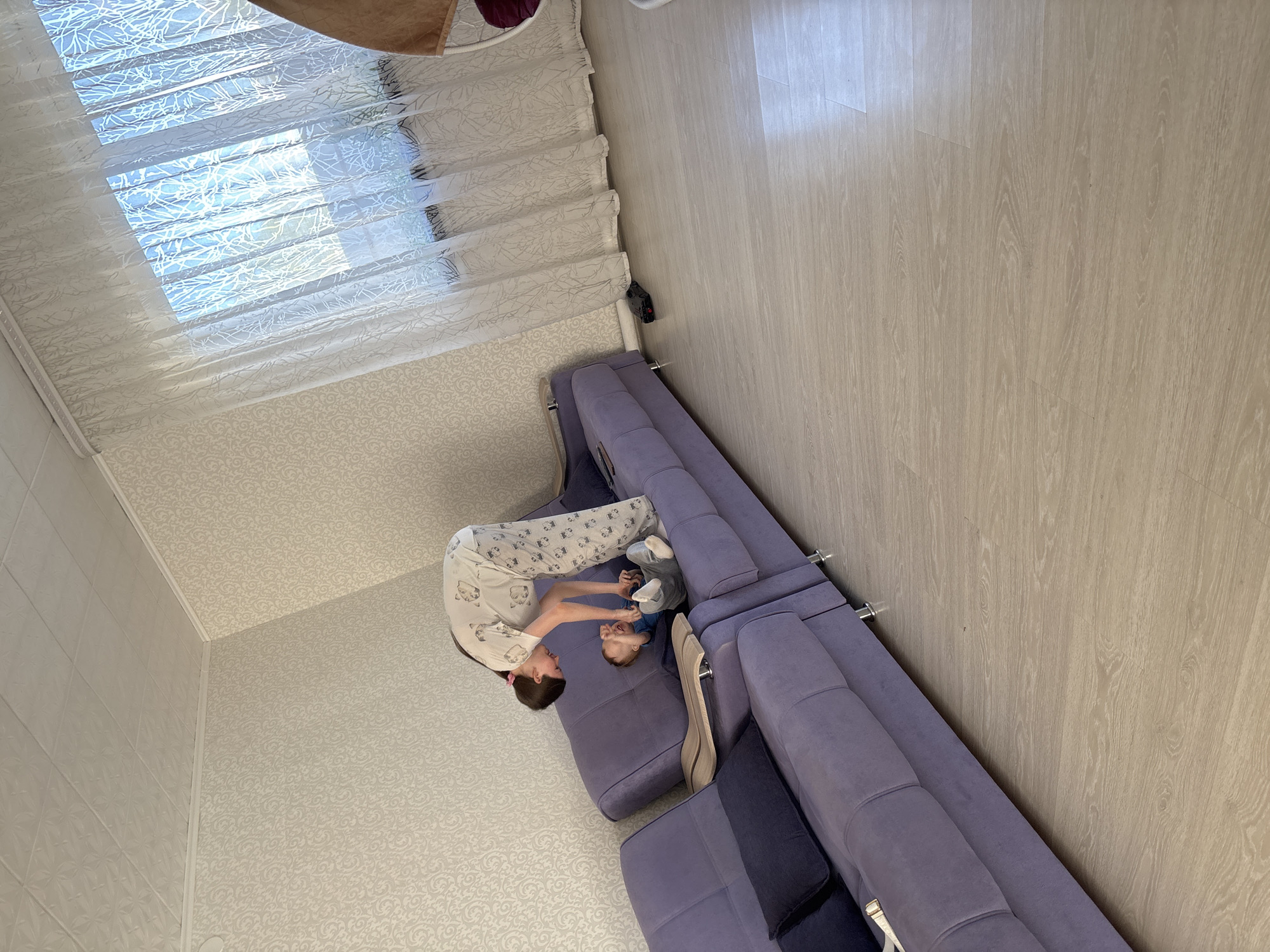 A high-angle shot reveals a domestic scene. A person in patterned pajamas interacts with a young child on a purple sectional sofa. The room is characterized by light-colored wood-grain flooring and textured wallpaper. Natural light filters through a window covered with sheer patterned curtains. The location is identified as Ферма, Russia.