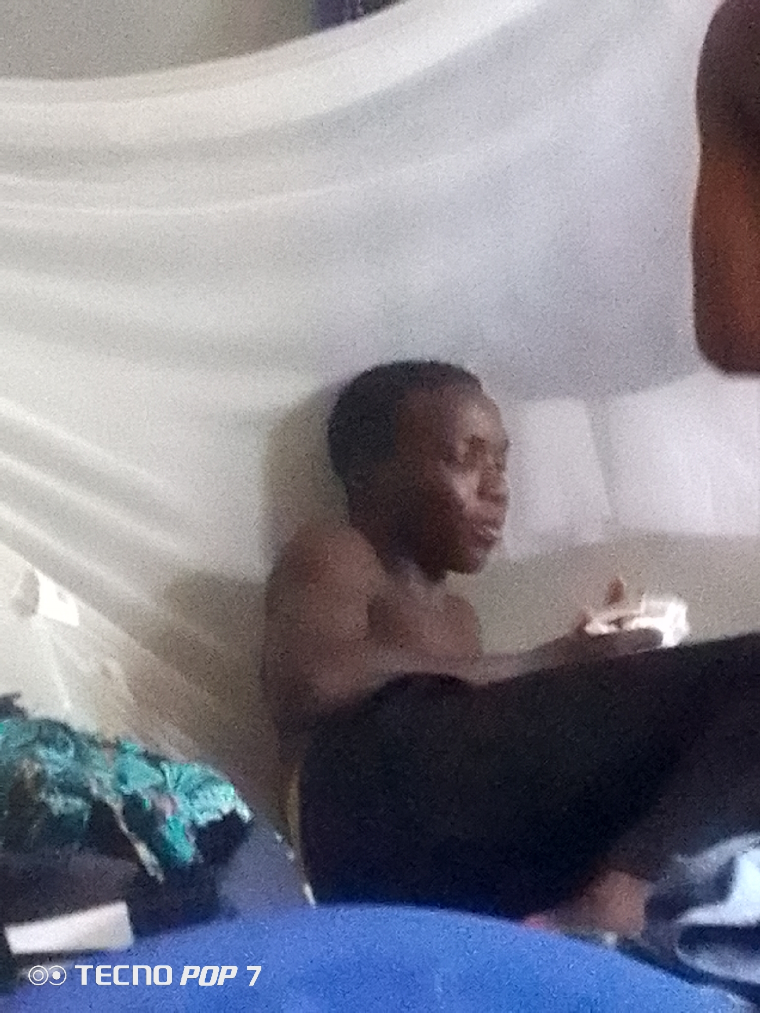 The image shows a close-up, low-light, candid shot of a person from the chest up, likely in a domestic setting. The person, appearing to be young with dark skin and short hair, is shirtless and looking slightly off-camera to their right, with their mouth slightly open as if speaking or about to speak. Their right hand is visible, holding a small, white object.
The background is dominated by a draped, white material that creates a soft, tent-like effect, possibly a mosquito net or a curtain. The lighting is dim, with some shadows and a grainy texture throughout the image. To the left of the person, there is a glimpse of colorful fabric, possibly clothing or a blanket. The overall scene suggests an informal, indoor environment, perhaps a bedroom or a living space.
Visible text at the bottom left corner of the image reads "TECNO POP 7" with a stylized logo, indicating the brand of the mobile device used to capture the photograph. There are no clear indicators of the time of day or weather, but the dim lighting suggests it could be evening or night. The expression on the person's face is neutral to slightly engaged.