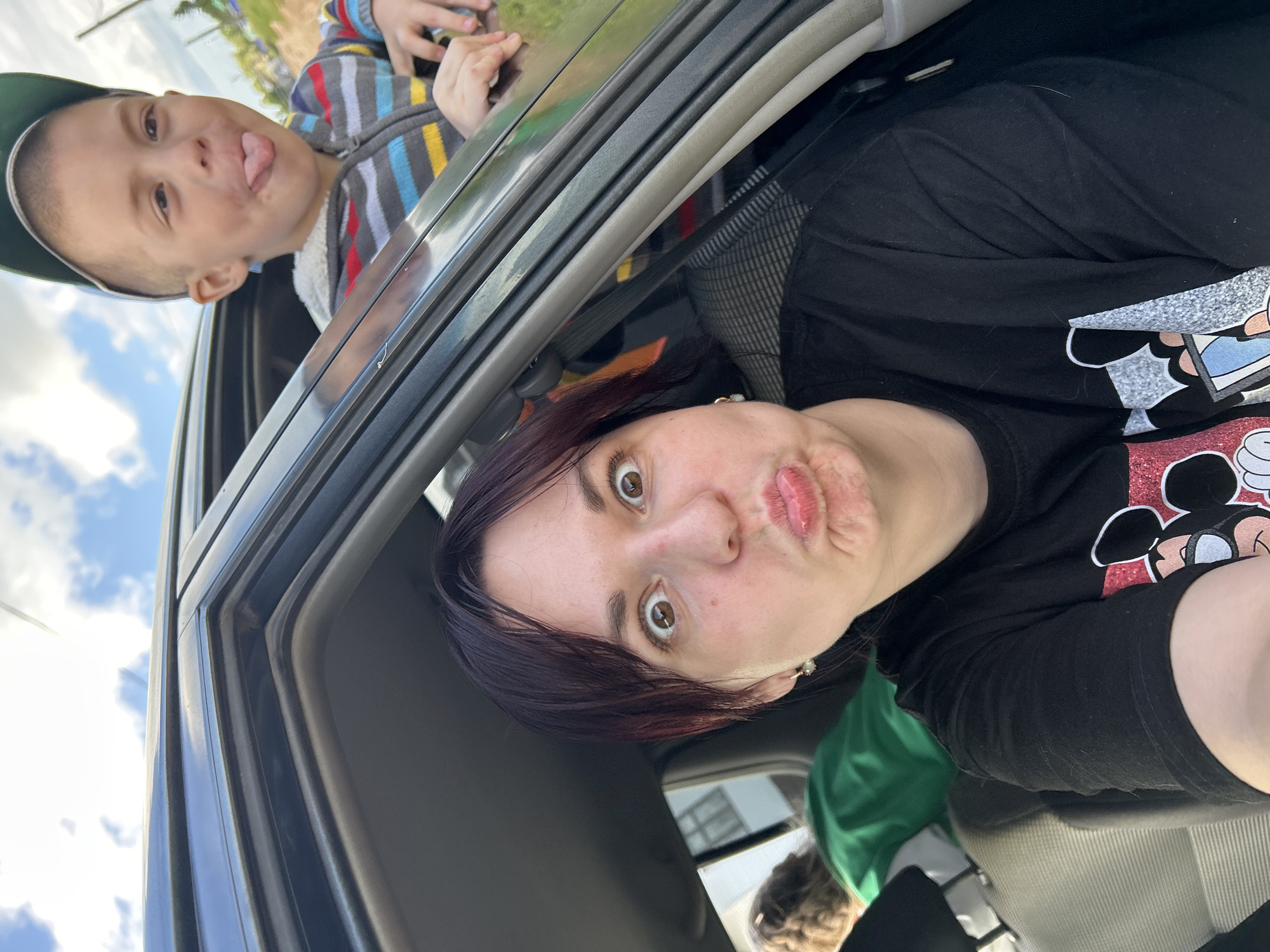 The image is a close-up, selfie-style shot taken from inside a car. A woman with dark hair is in the foreground, making a duck face with her eyes wide open. She is wearing a black t-shirt with a Mickey Mouse graphic. To her right, and slightly behind her, a young boy is visible with his head and shoulders out of the car window. He is wearing a striped hoodie and a baseball cap, and is sticking his tongue out. Another person is visible in the back seat, wearing a green top. The background shows a cloudy sky and a grassy area, suggesting an outdoor, possibly rural, setting. The specific location in the city of Fokino, Russia, cannot be determined from the visual information provided.