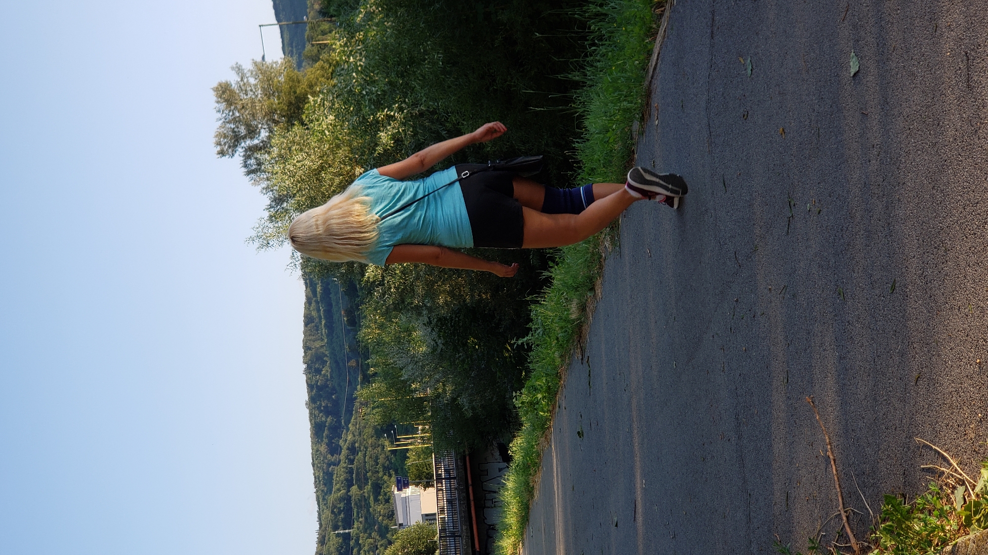 The image shows a woman from behind, walking on a paved path next to a grassy area. She has blonde hair and is wearing a turquoise t-shirt, black shorts, a blue knee brace, and sneakers. She is carrying a black bag over her right shoulder. In the background, there are trees, a bridge, and buildings. The scene appears to be in a park or recreational area. The lighting suggests it is either early morning or late afternoon. The location is indicated as being in the city of Prešov, Slovakia.