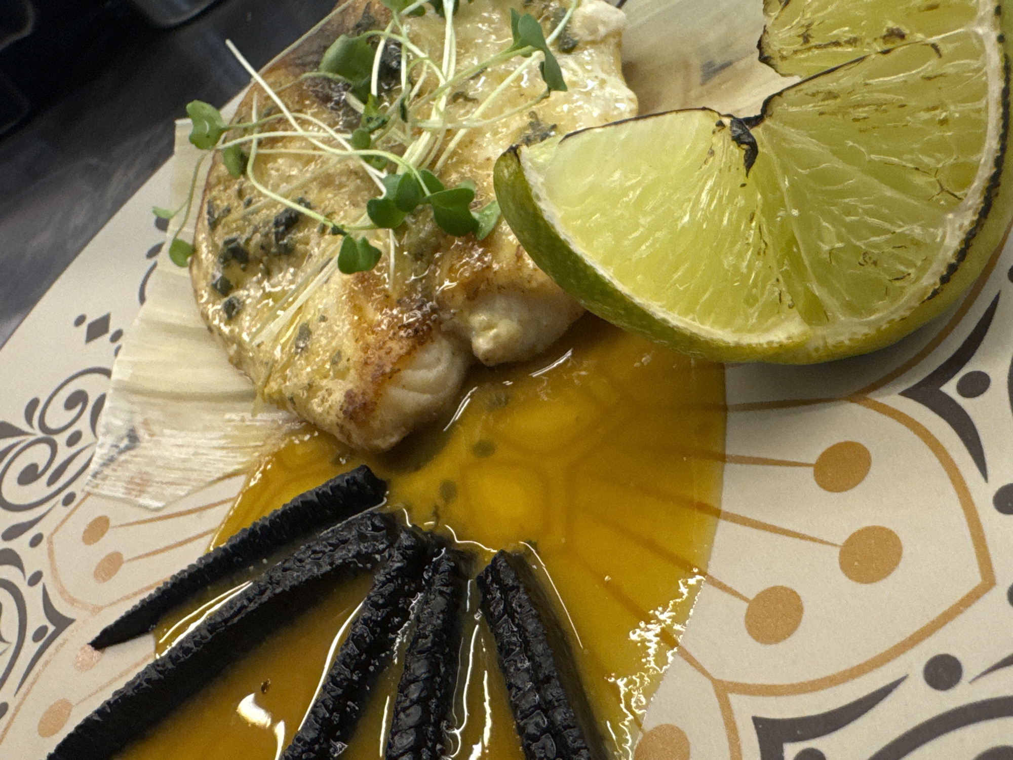 A close-up, overhead shot captures a gourmet dish served on a patterned plate, possibly in a restaurant setting. The plate, a light cream color, features a geometric design with brown and grey swirling lines, punctuated by clusters of three small, light brown circles. A vibrant, golden-orange sauce fills a significant portion of the plate's left side. Arranged in the sauce are three dark, almost black, elongated pieces of food, possibly charred vegetables or a type of crispy bread, that appear to be cut into uniform segments.
Dominating the right side of the frame is a piece of cooked white fish, glistening with sauce and herbs, possibly rosemary, which has been pan-seared to a golden brown. It's garnished with delicate, pale green microgreens. Partially obscuring the fish and sauce is a large wedge of bright green lime, with its textured rind and juicy, segmented interior. The lime appears to have been lightly grilled or seared, as its edges are slightly darkened.
The lighting is warm and focused, highlighting the textures and colors of the food. The background is blurred, suggesting a shallow depth of field. There is no visible indication of the city of Sofia, Bulgaria, or any specific geographical location. The scene is entirely focused on the presentation and composition of the food.