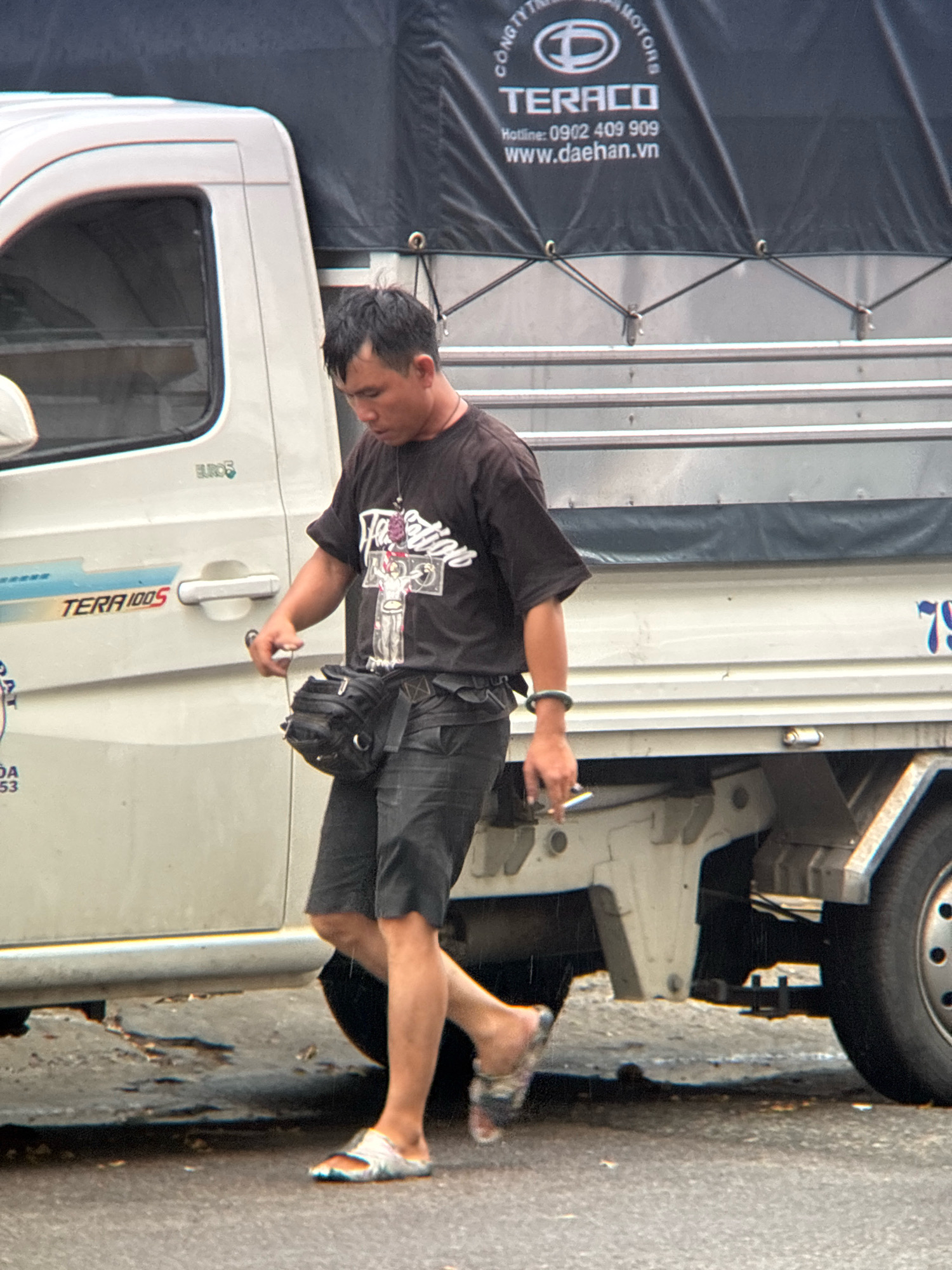 A man in a black t-shirt and dark shorts stands beside a white truck in Nha Trang, Vietnam. The truck has a black tarp covering its cargo area with the logo "TERACO" visible. The man is wearing a black fanny pack around his waist and flip-flops. He appears to be holding a cigarette in his right hand. The ground is wet, suggesting recent rain. The scene depicts a street or roadside environment.