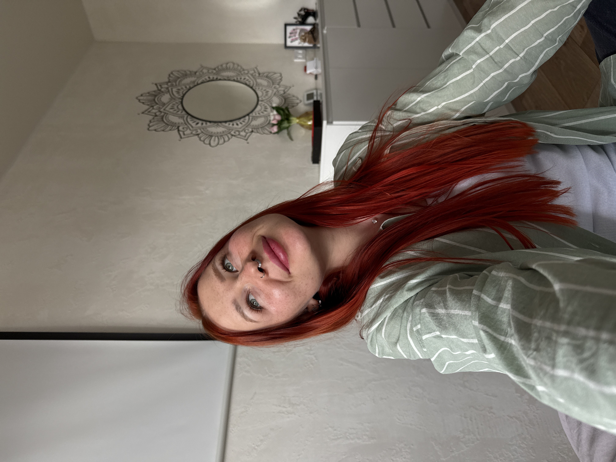 An upward-facing selfie captures a young woman with vibrant, long red hair and light eyes, wearing a light green and white striped shirt. She has a subtle smile and freckles on her nose and cheeks, and a silver septum piercing. Her gaze is directed towards the camera.
The setting appears to be an indoor room, likely a home in Rostov-on-Don, Russia. Behind her, a light-colored, textured wall features a decorative round mirror with an ornate, mandala-like frame. Below the mirror, a golden vase holding pink flowers (possibly tulips) is visible on a surface, alongside a small framed picture and other indistinct objects on shelves in the background. To the far left, a large white rectangular screen, possibly a projector screen or blind, is partially visible. The woman's attire also includes a lighter garment underneath her striped shirt and dark blue fabric (likely jeans) to the far right of the frame. The lighting suggests it is daytime. No visible text is present in the image.
